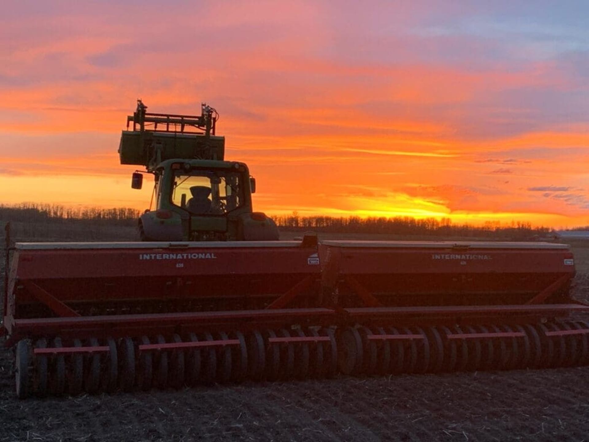 Tractor with large red seeder in field under bright orange sunset sky.