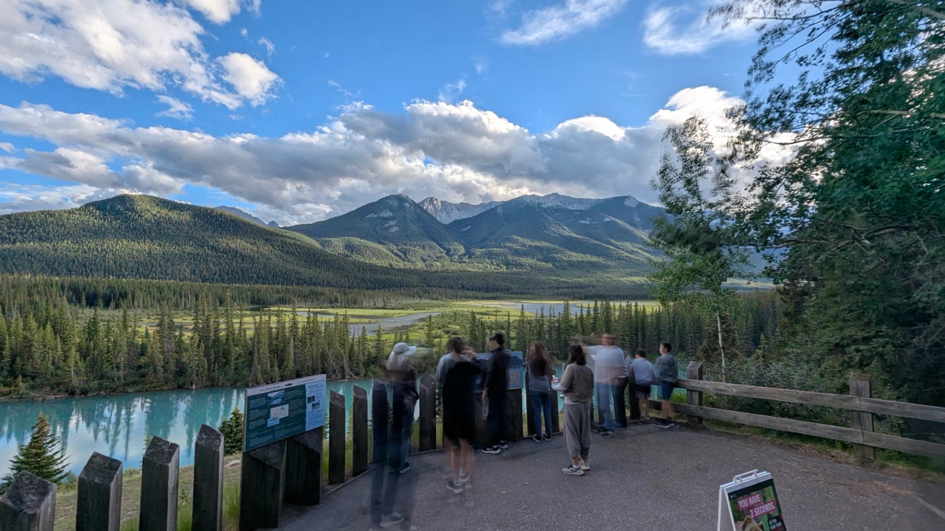 Visitors overlook a turquoise river, evergreen forest and mountains beneath a cloud-filled sky.