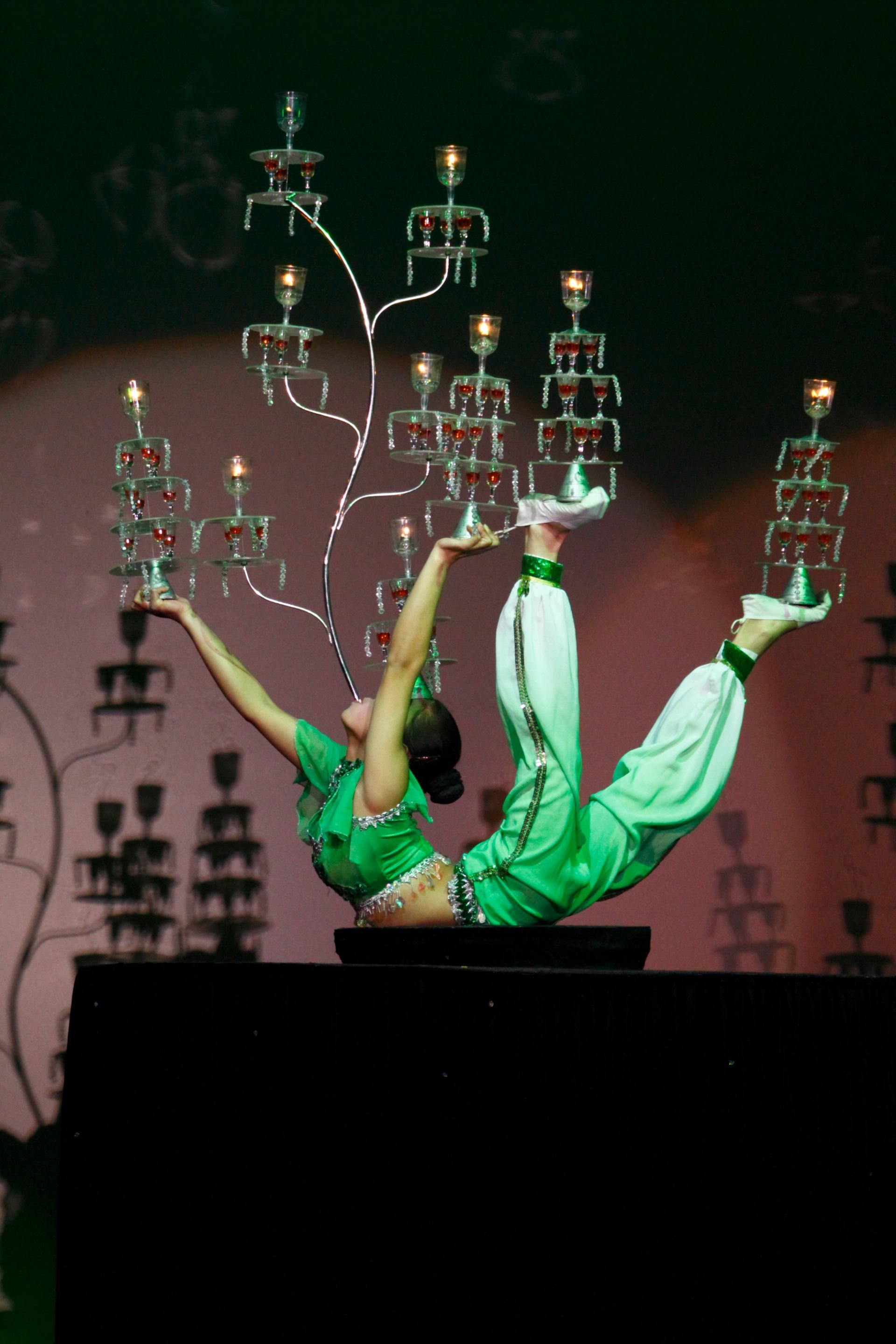 Acrobat in green costume balances stacked glass candelabras while bending backward on platform.