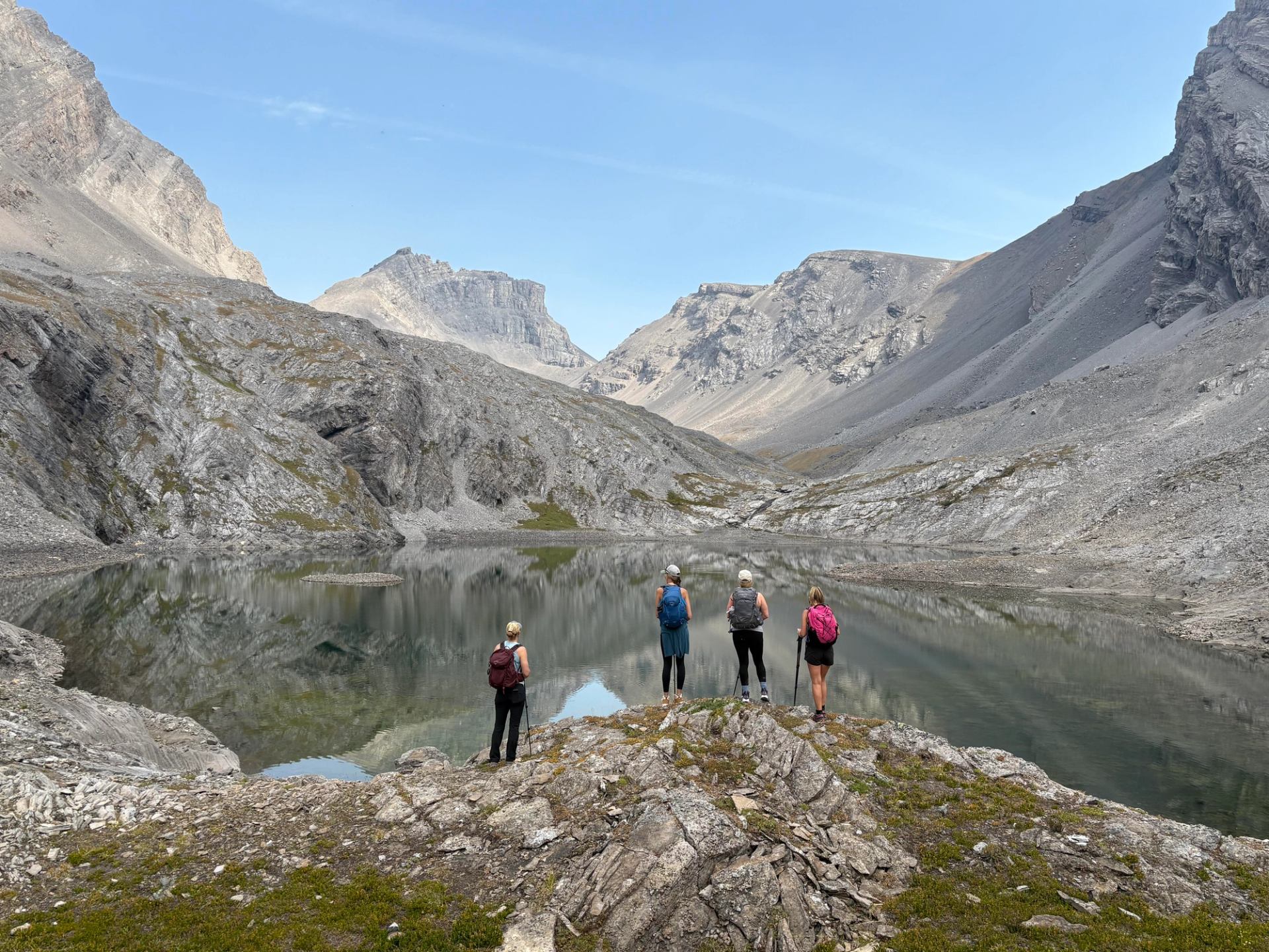 Group of hikers standing by a reflective alpine lake surrounded by rocky peaks.