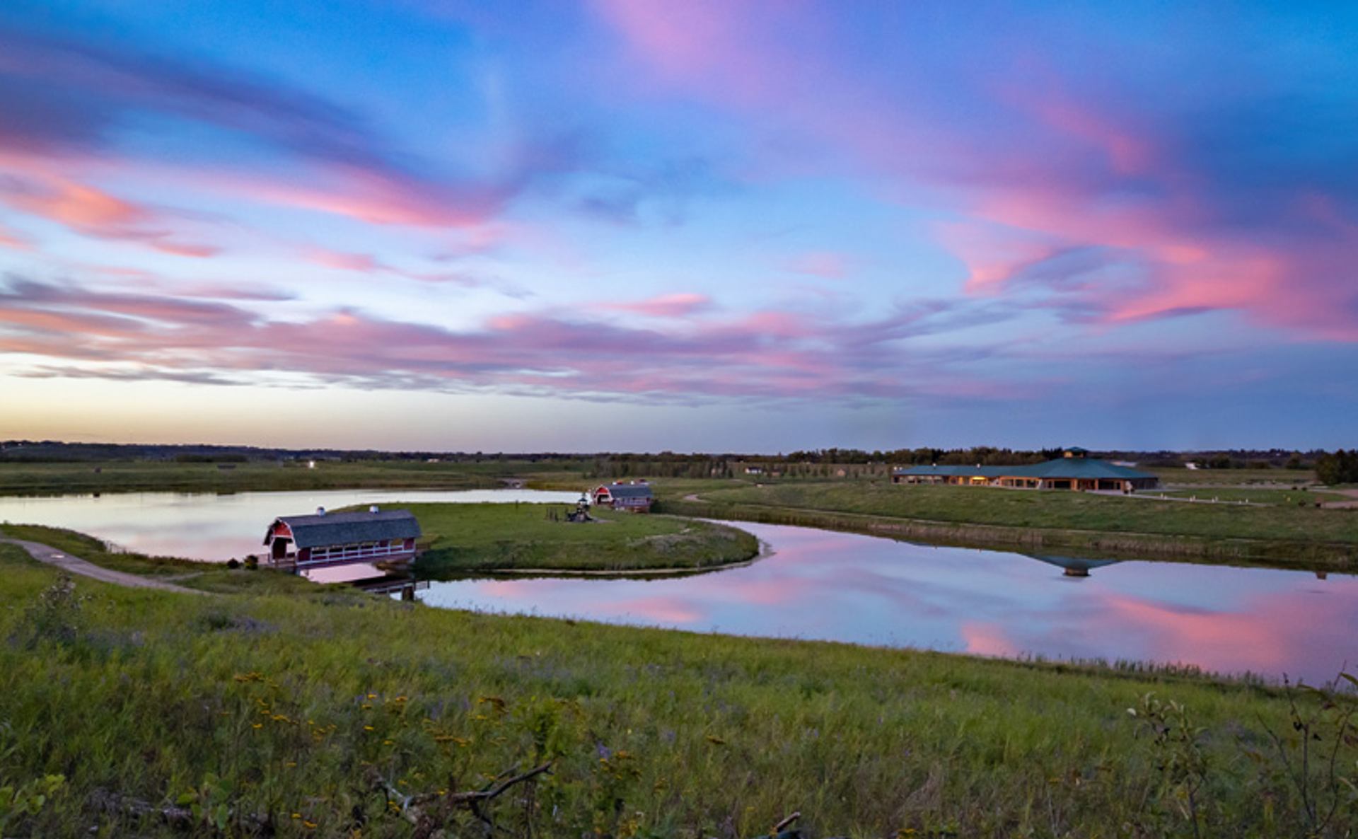 Small barn beside winding pond under pink and blue clouds at dusk.