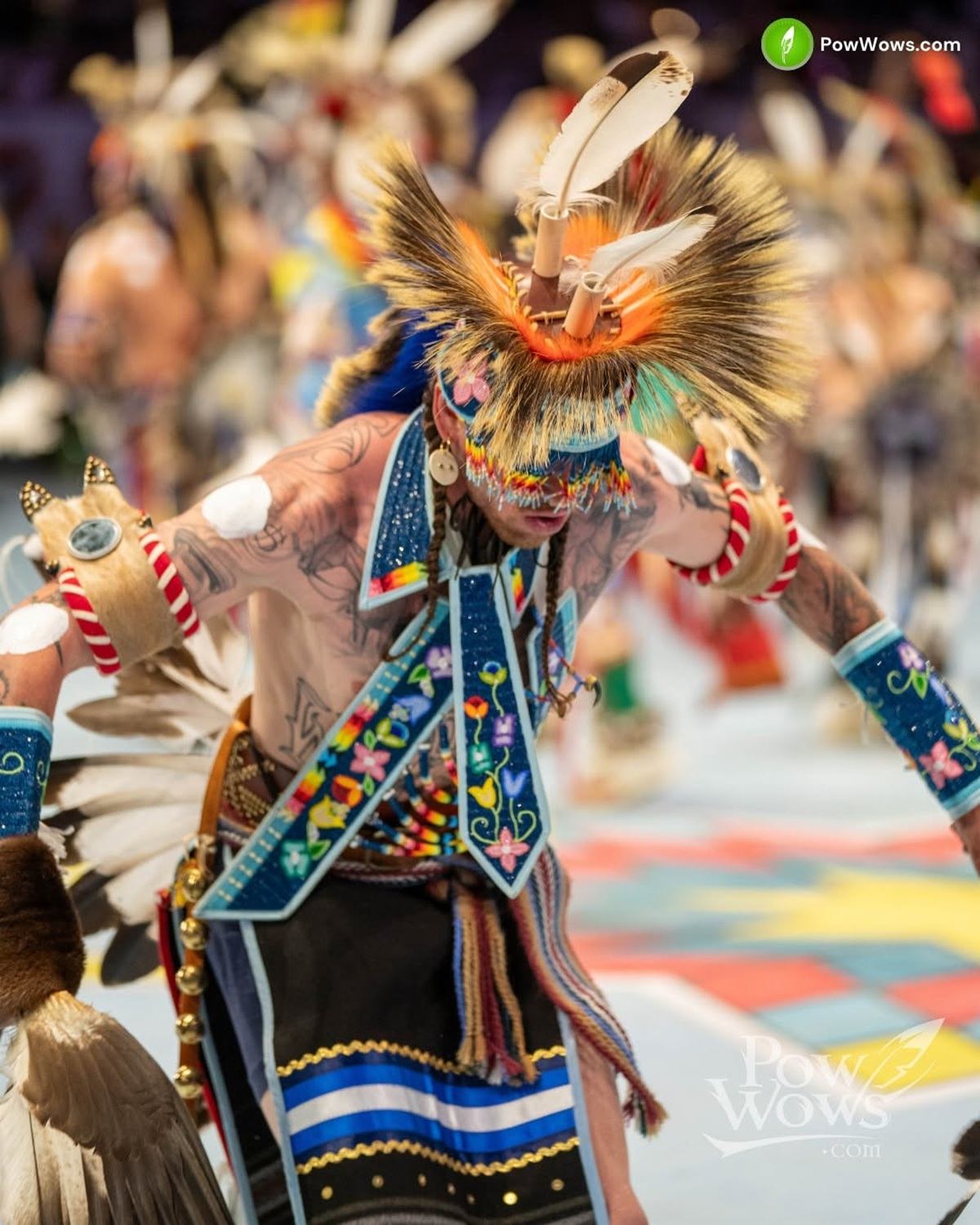 Powwow dancer in colourful regalia with feathered headdress performs on patterned arena floor.