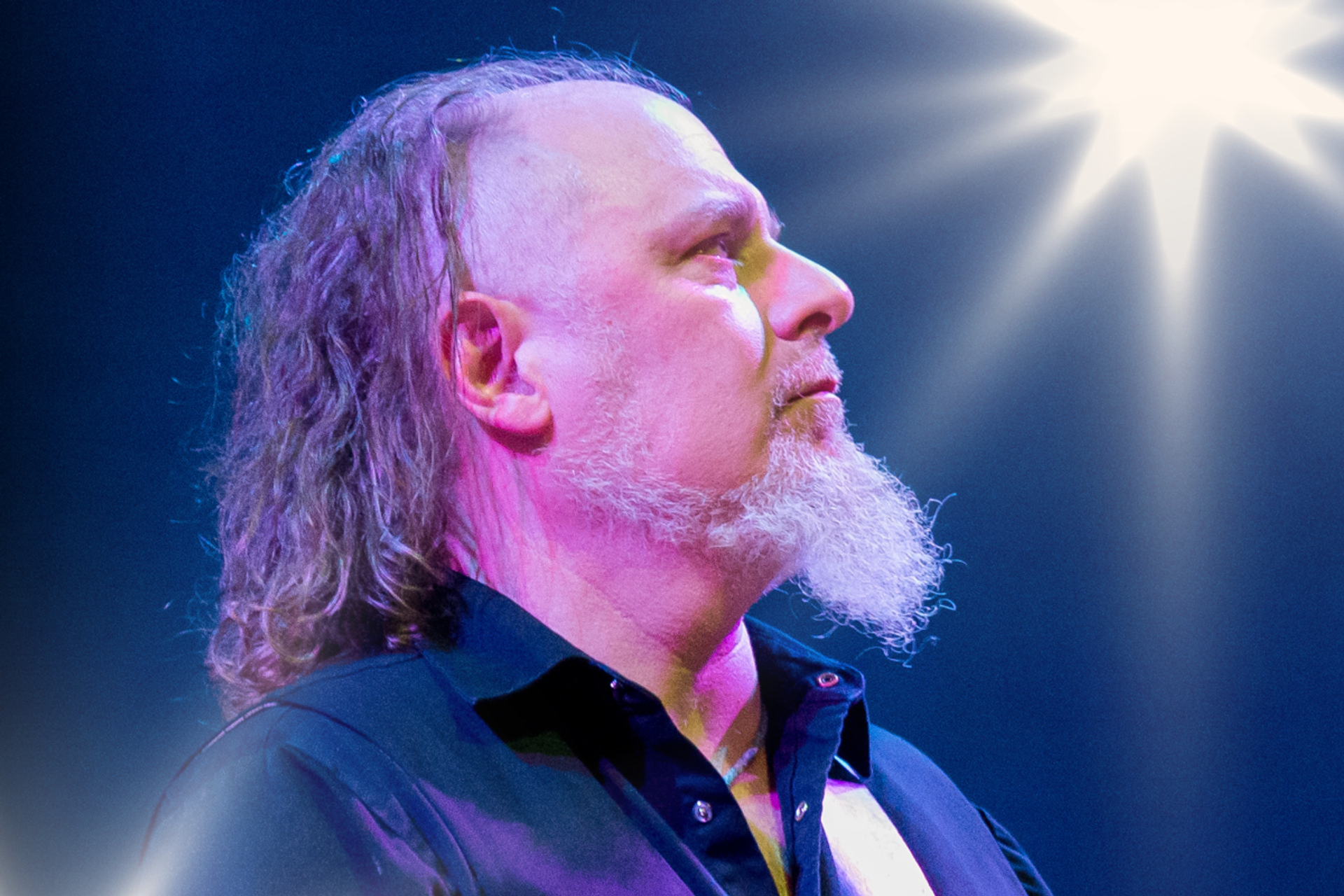 Musician with long hair and white beard onstage under bright concert lights.