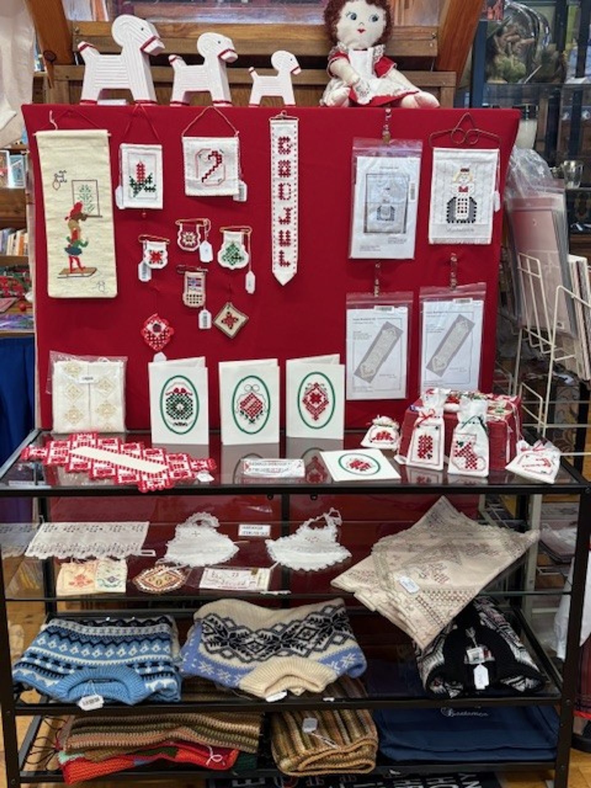 A retail display of traditional Scandinavian crafts, including embroidered textiles, cross-stitch patterns, wooden horse figures, and knitted sweaters.