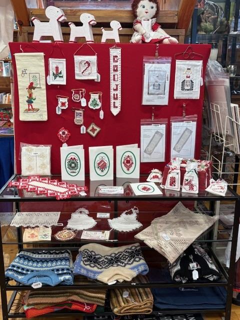 A retail display of traditional Scandinavian crafts, including embroidered textiles, cross-stitch patterns, wooden horse figures, and knitted sweaters.