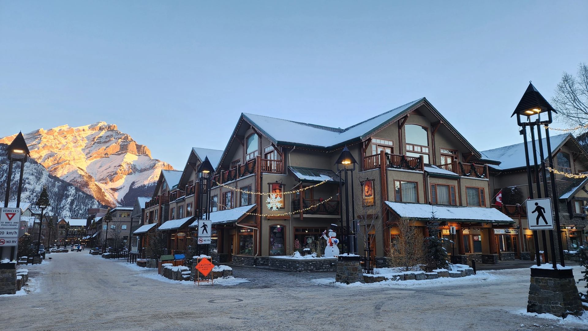 Rustic lodge with festive lights in snowy mountain setting under a sunlit peak.