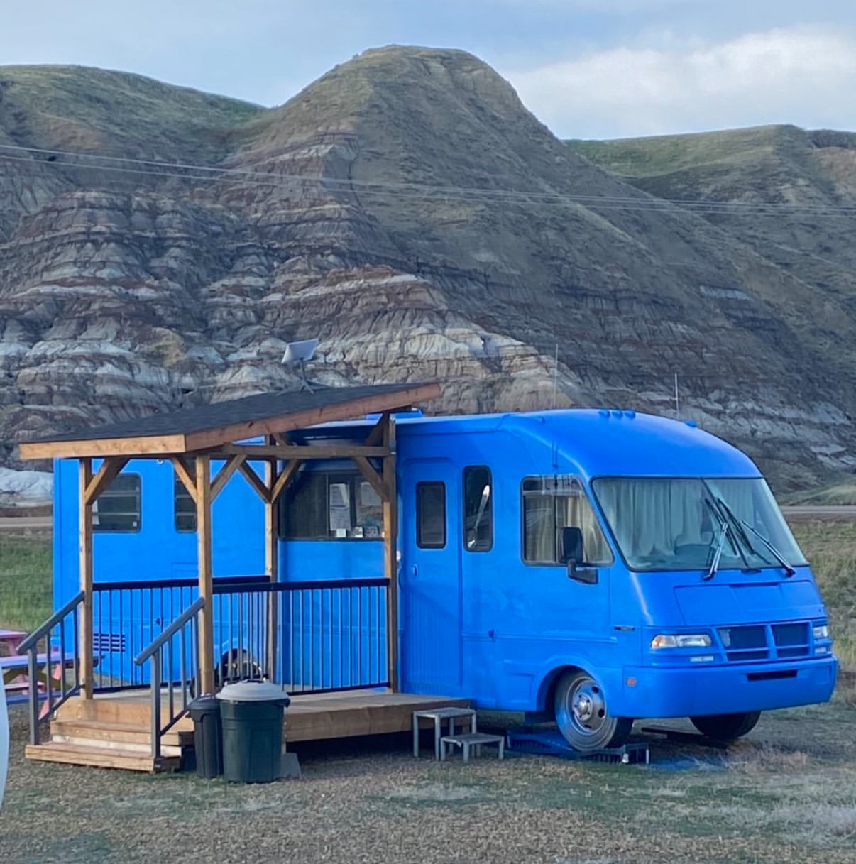 Blue RV with attached wooden deck in scenic outdoor setting with hills in background.