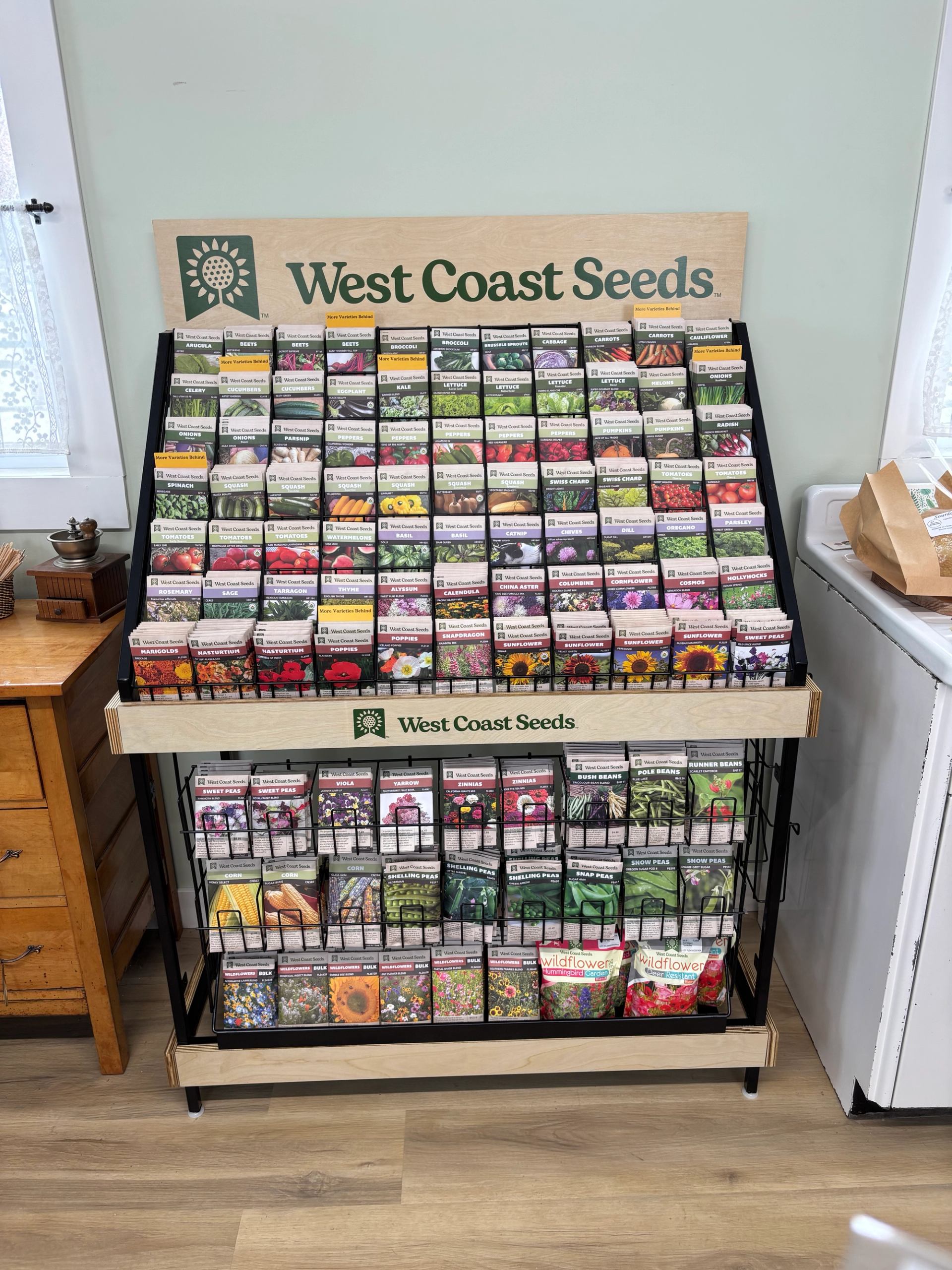 A large wooden and metal display stand featuring hundreds of West Coast Seeds packets for various vegetables, fruits, and flowers.