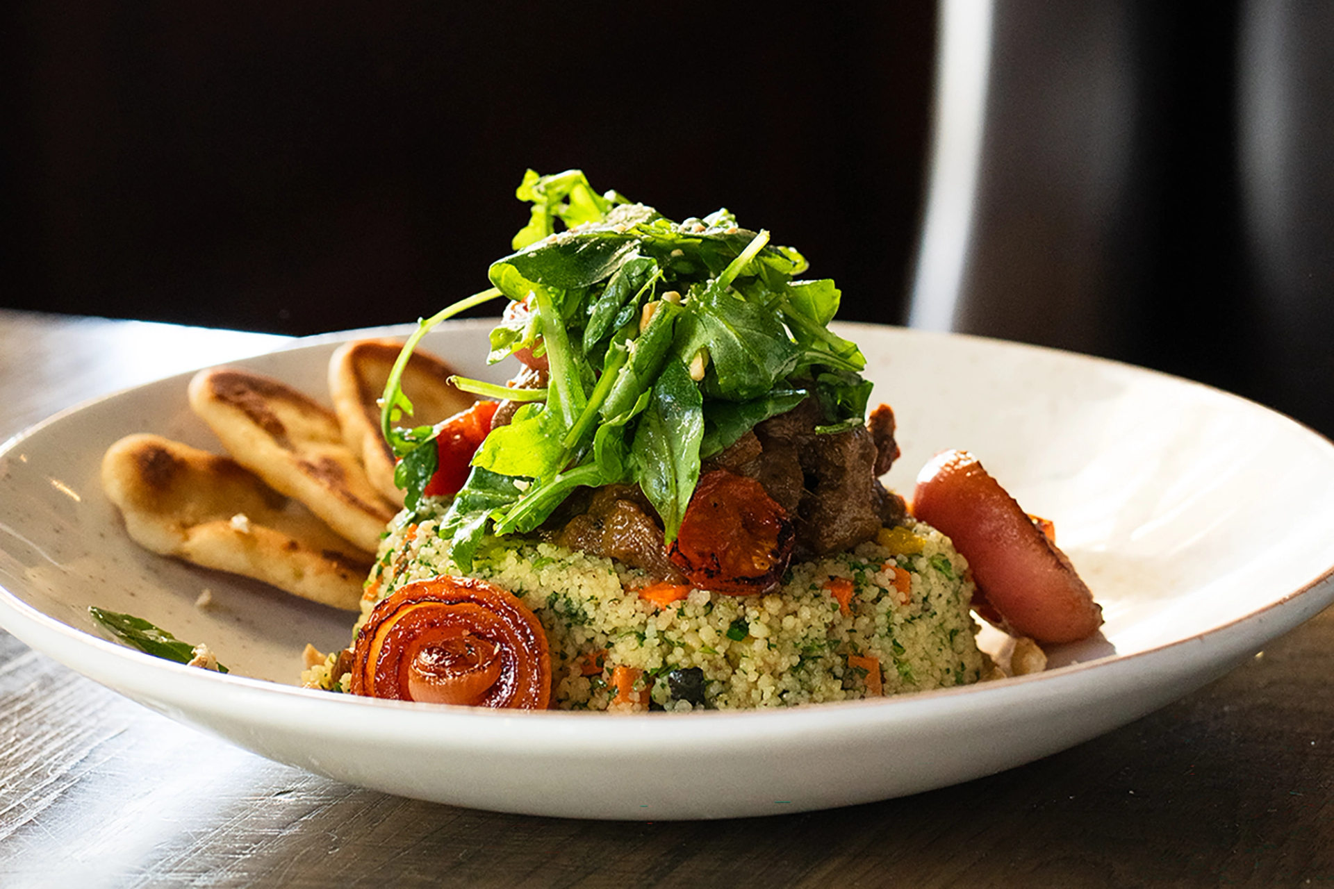 Plate of couscous with roasted vegetables, leafy greens and flatbread on wooden table.