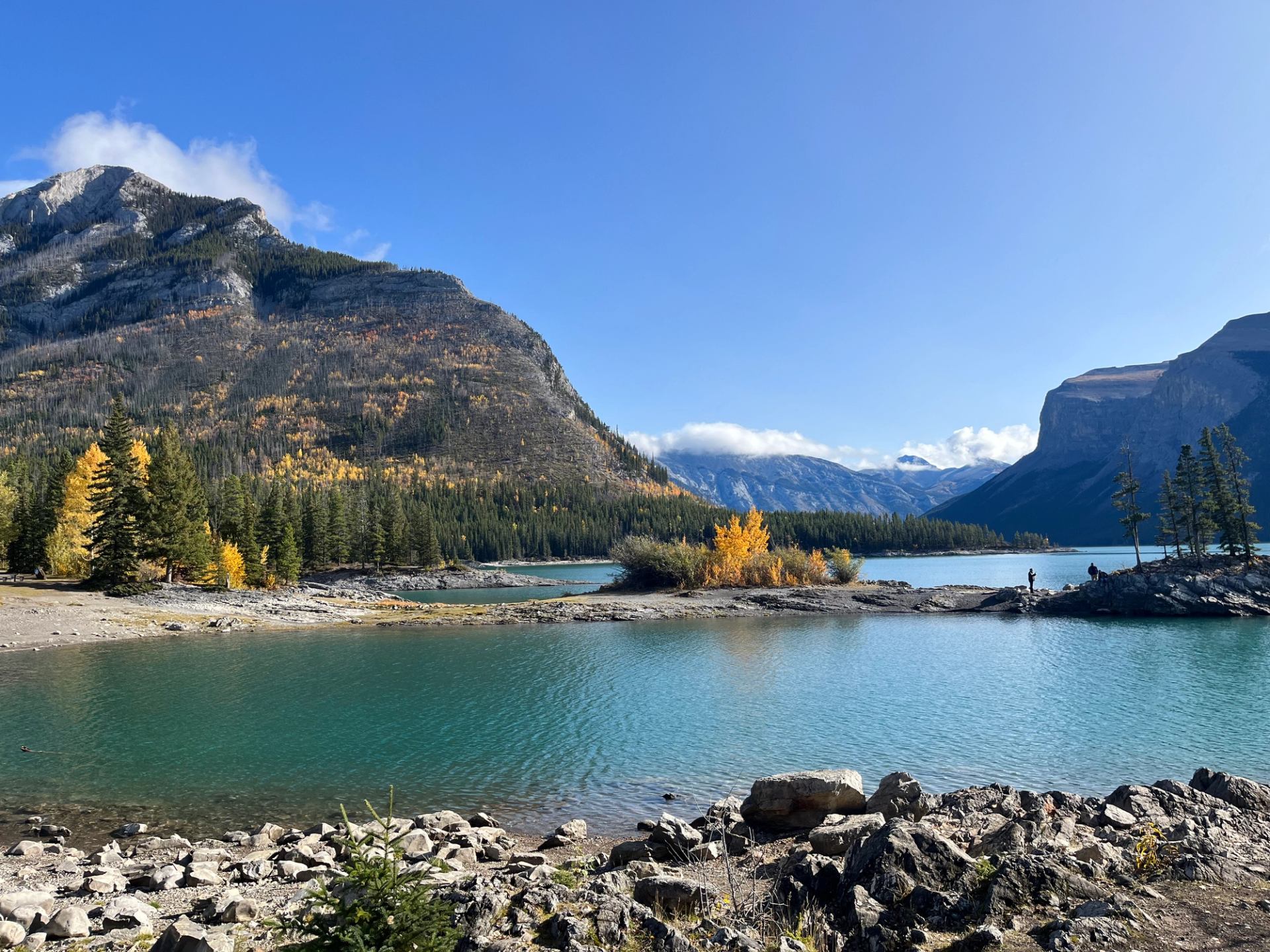 Mountain lake with turquoise water, rocky shore, evergreen trees and distant hikers.