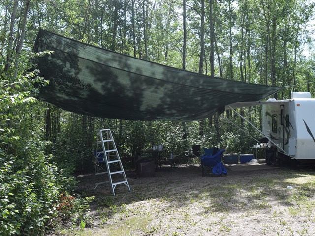 Tarp shelter set up beside an RV in a wooded campsite with chairs and a ladder.
