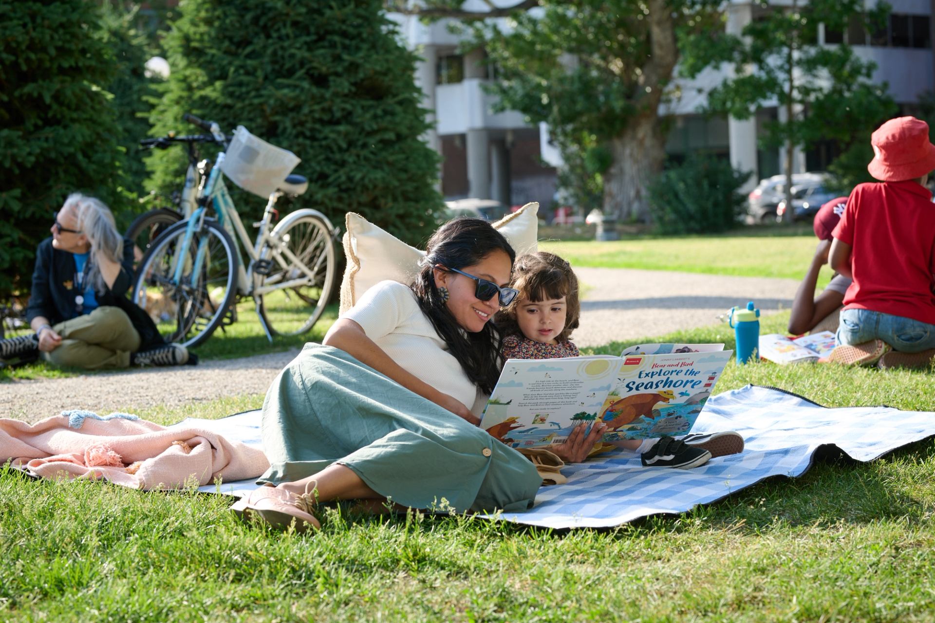Adults and children read picture books on blankets in a sunny park.