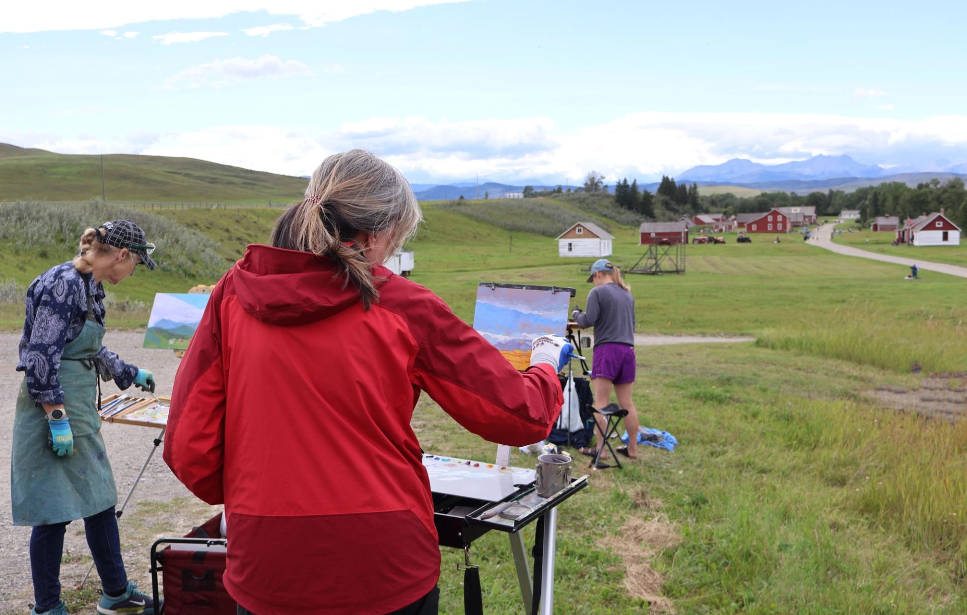 Artists paint on easels outdoors overlooking rolling hills and historic ranch buildings.