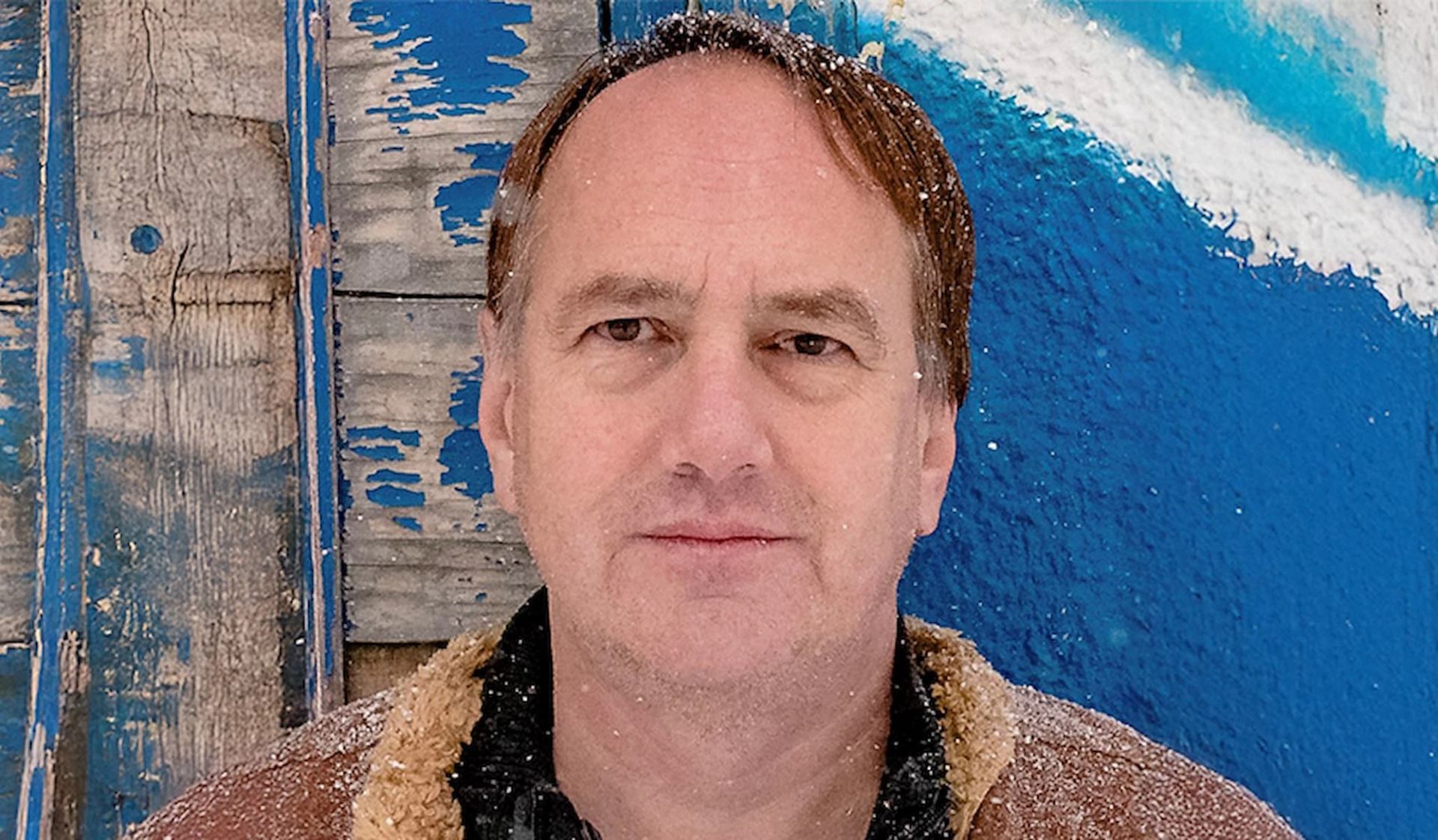 Person with snow on hair and coat stands before weathered blue wooden wall.