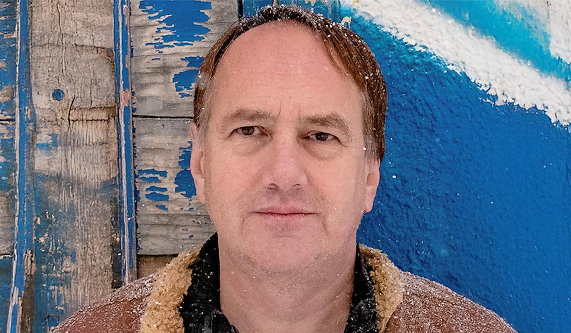 Person with snow on hair and coat stands before weathered blue wooden wall.