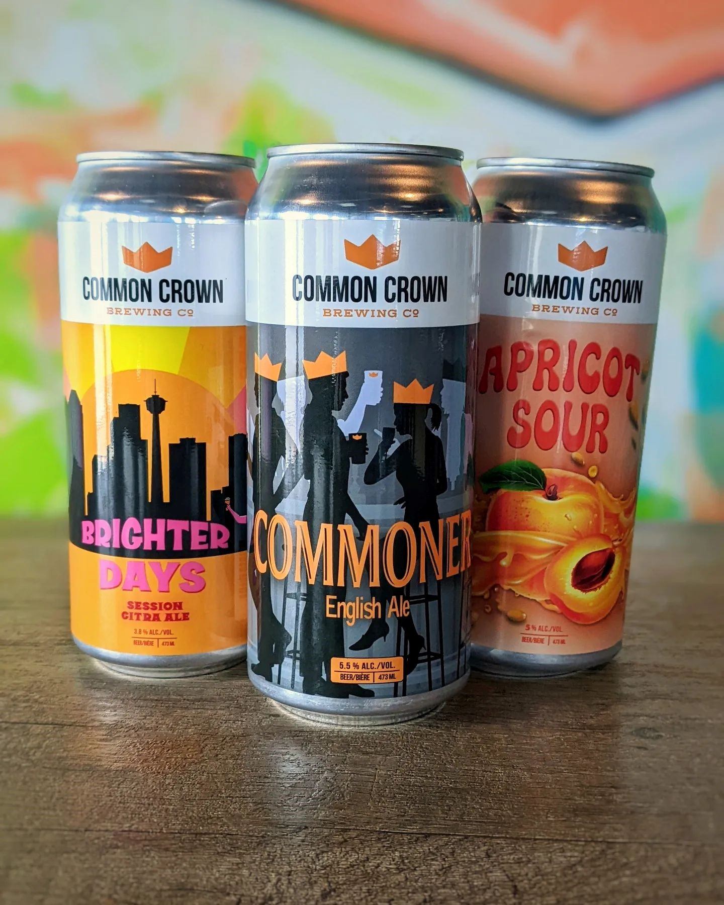 Three Common Crown beer cans on wood surface with colorful background.