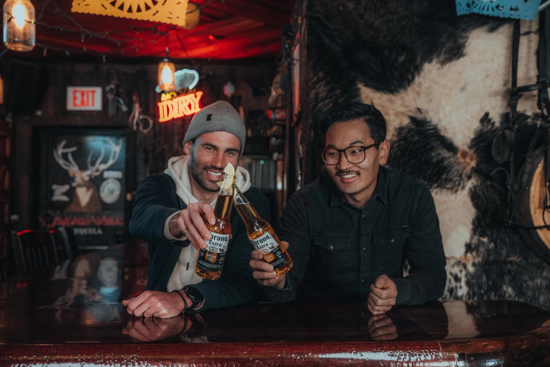 Two men smile while clinking Corona beer bottles at a bar.