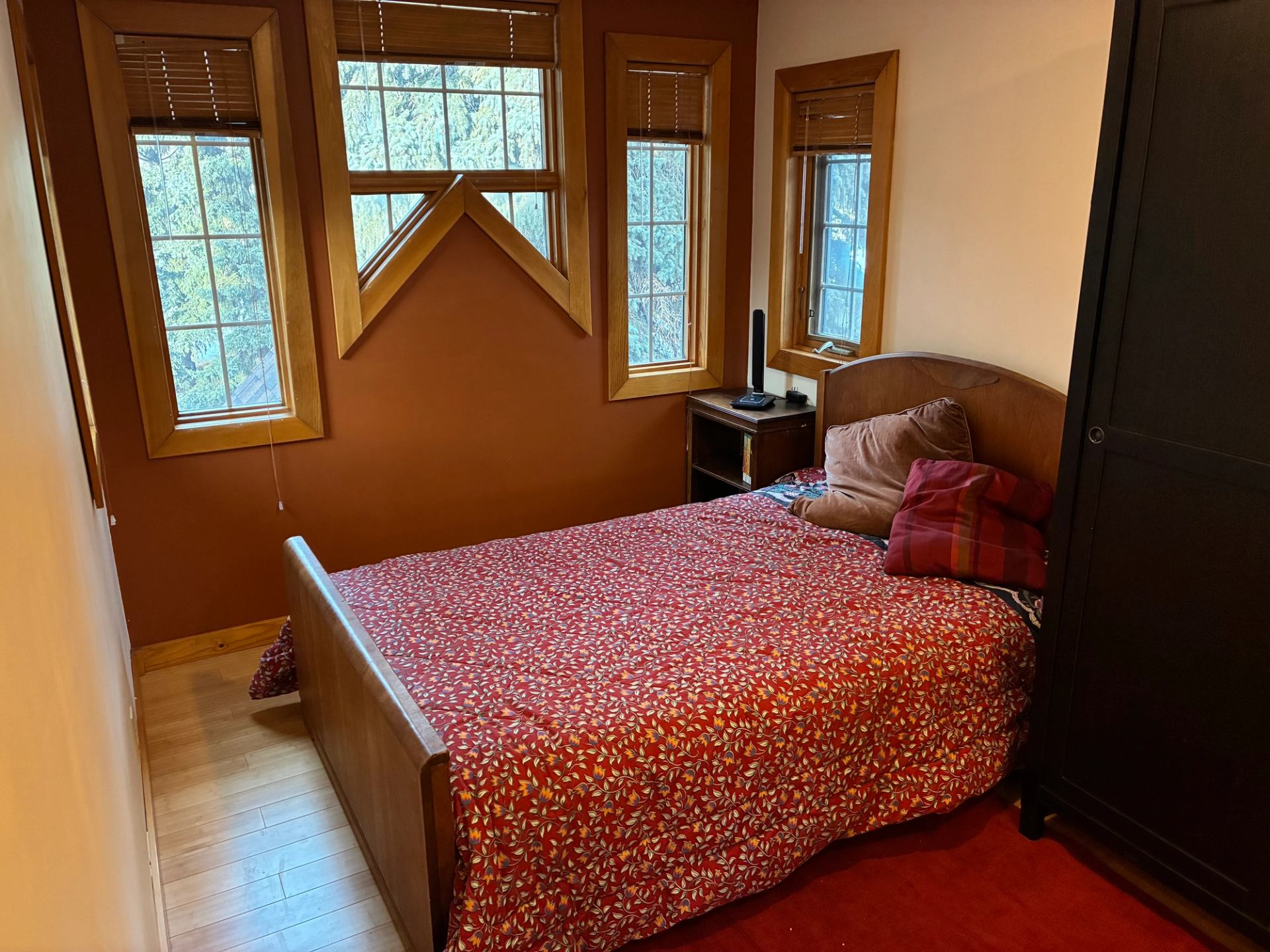 Small bedroom with red patterned bedspread, wooden windows, nightstand and dark wardrobe.