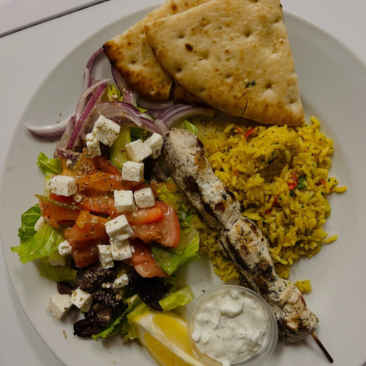 Plate with chicken skewer, yellow rice, Greek salad, pita bread, and tzatziki