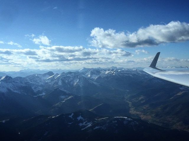 View from a glider soaring above rugged snow-capped mountains under a bright blue sky.
