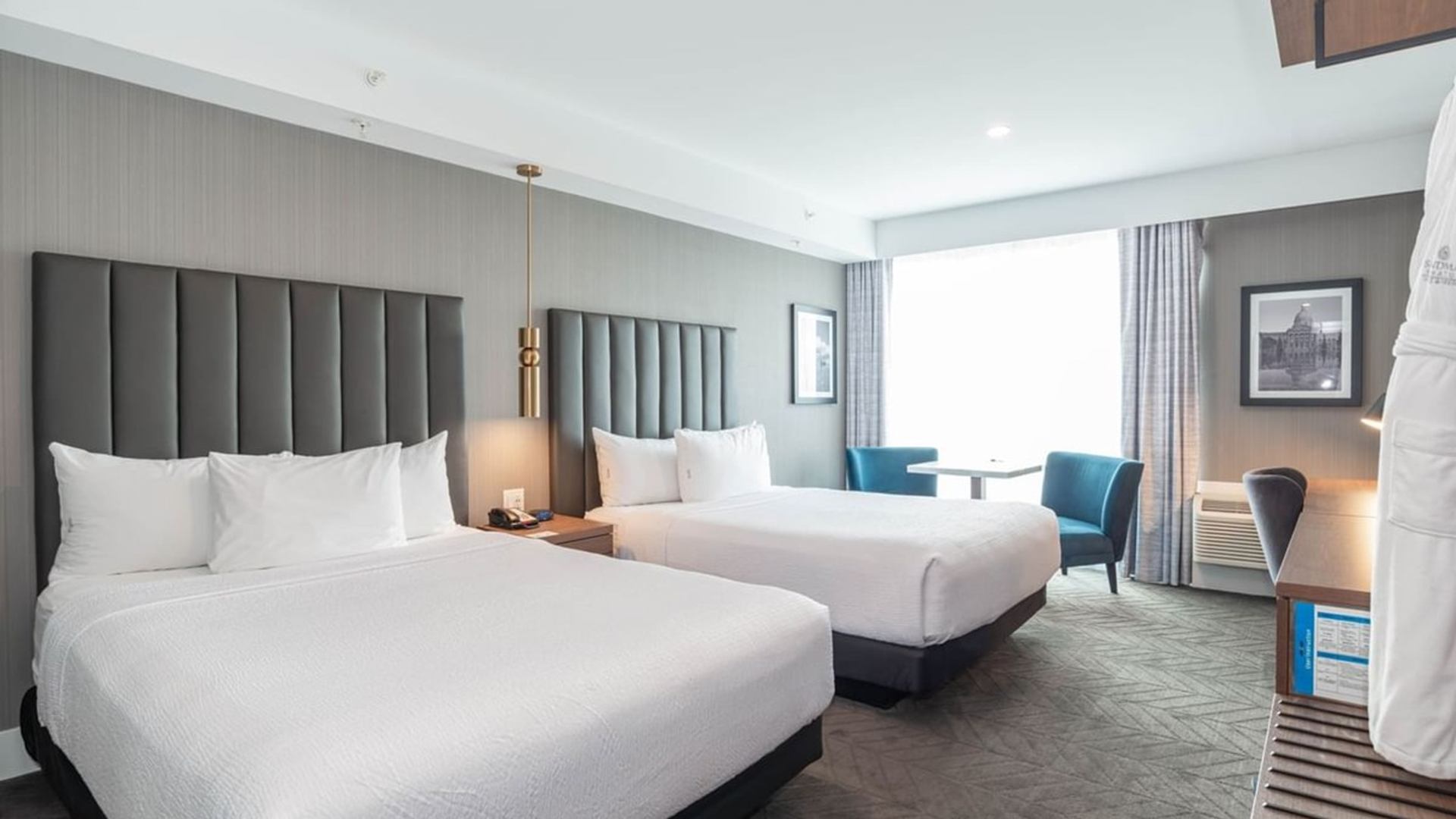Modern hotel room with two queen beds, upholstered headboards, bedside lighting, a window seating area with chairs and table, and contemporary decor.
