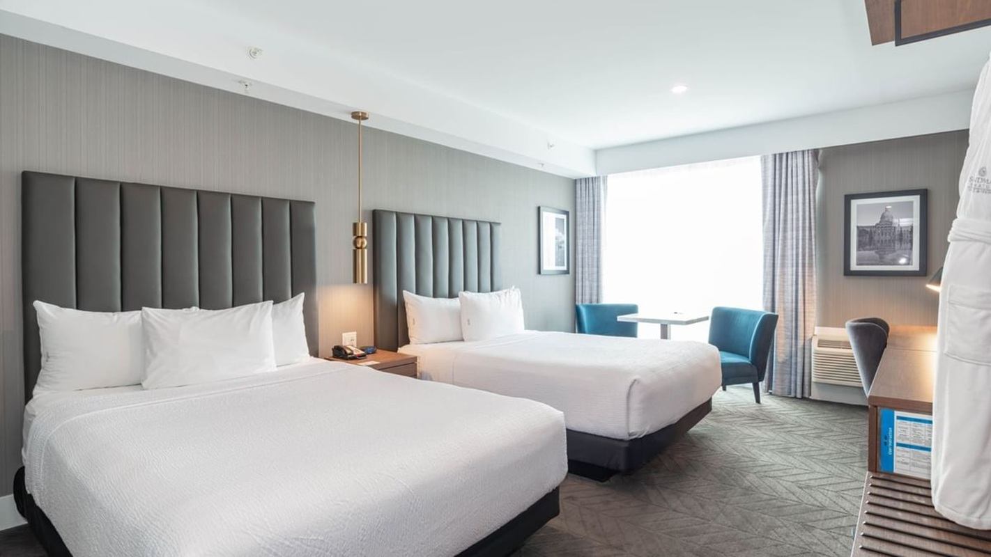 Modern hotel room with two queen beds, upholstered headboards, bedside lighting, a window seating area with chairs and table, and contemporary decor.