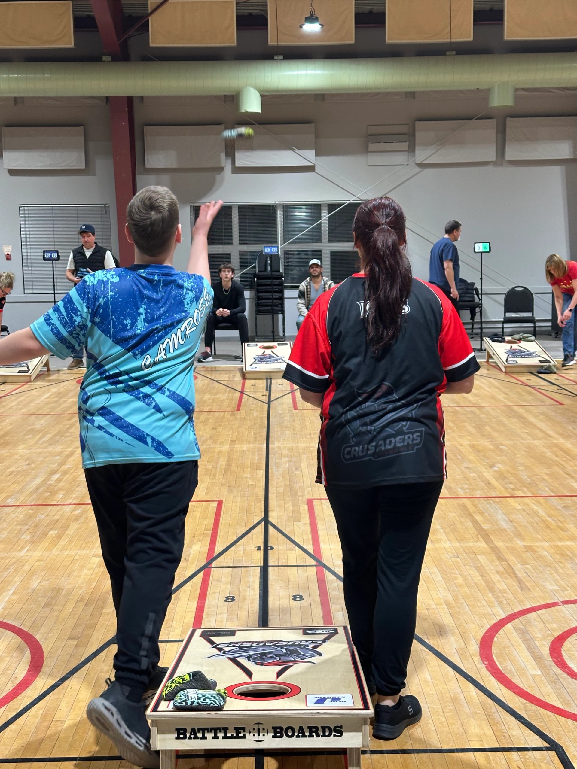 Two players tossing bags toward cornhole boards in a gym setting
