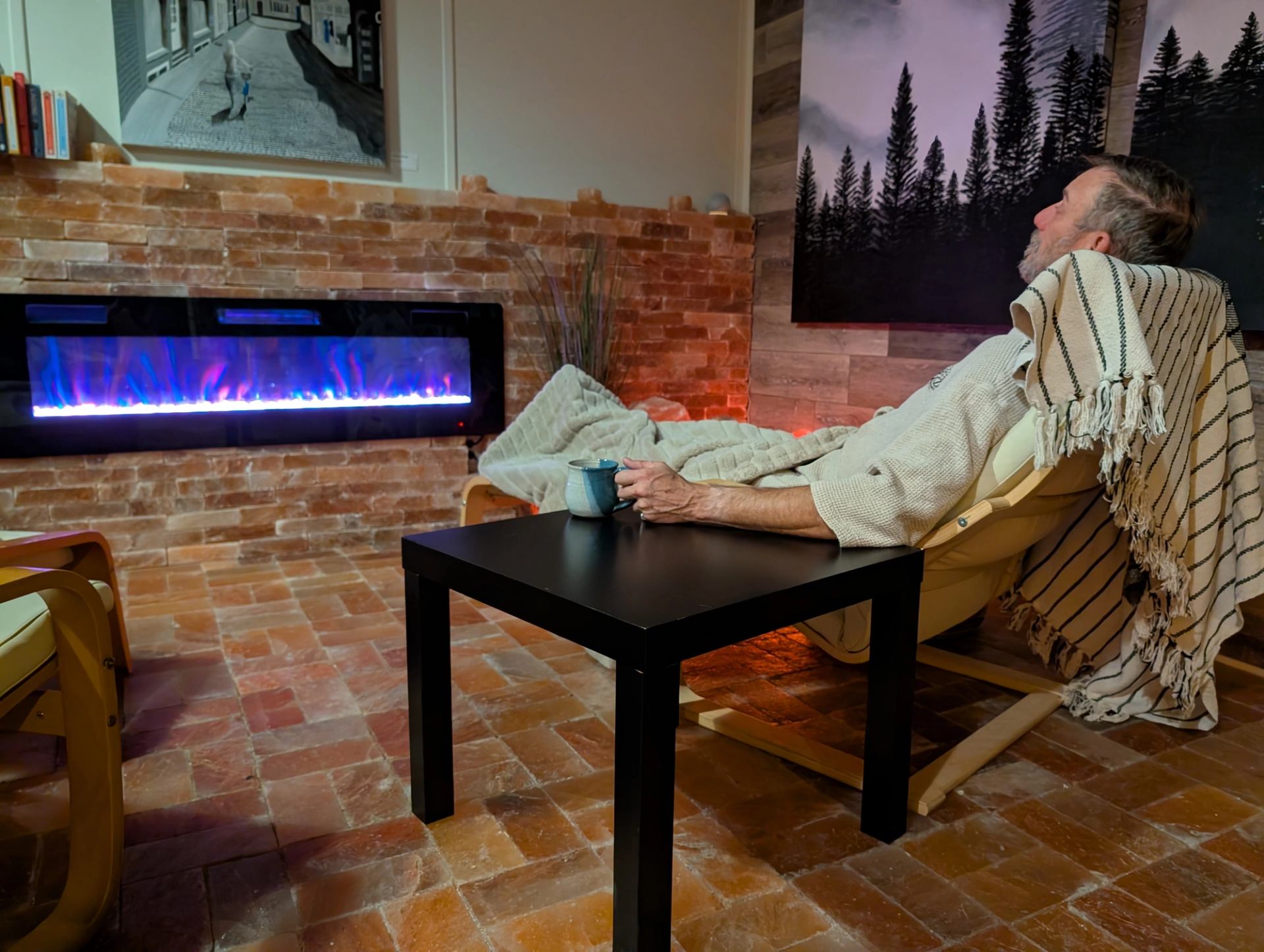 A man rests deeply in a comfortable lounge chair with his eyes closed, holding a warm mug. He is bundled up in a blanket in front of a colorful electric fireplace within the soothing salt relaxation room.