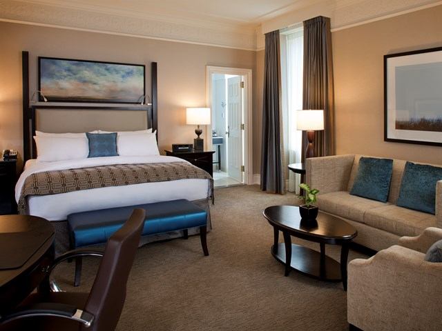 A well-lit hotel room features a large bed with pillows, a sofa, and a coffee table. Soft lighting and neutral tones create a cozy, inviting atmosphere.