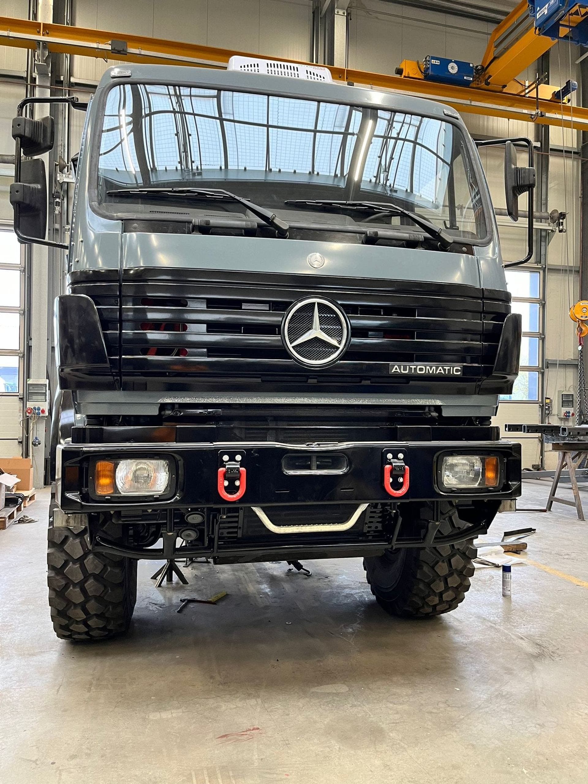 Black Mercedes-Benz expedition truck in garage with off-road tires and red tow hooks.