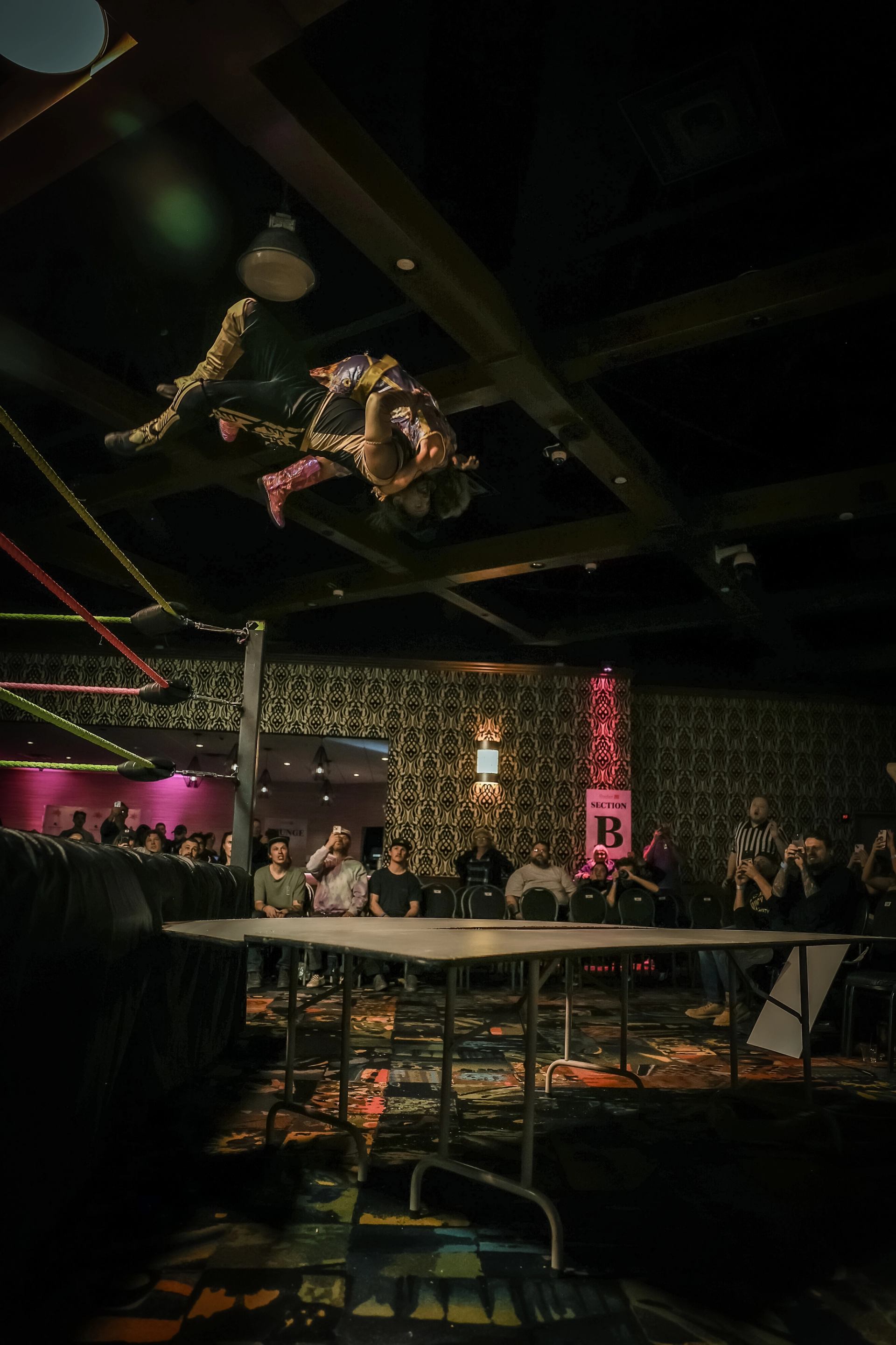 Two wrestlers flip through the air above a table with the crowd watching.