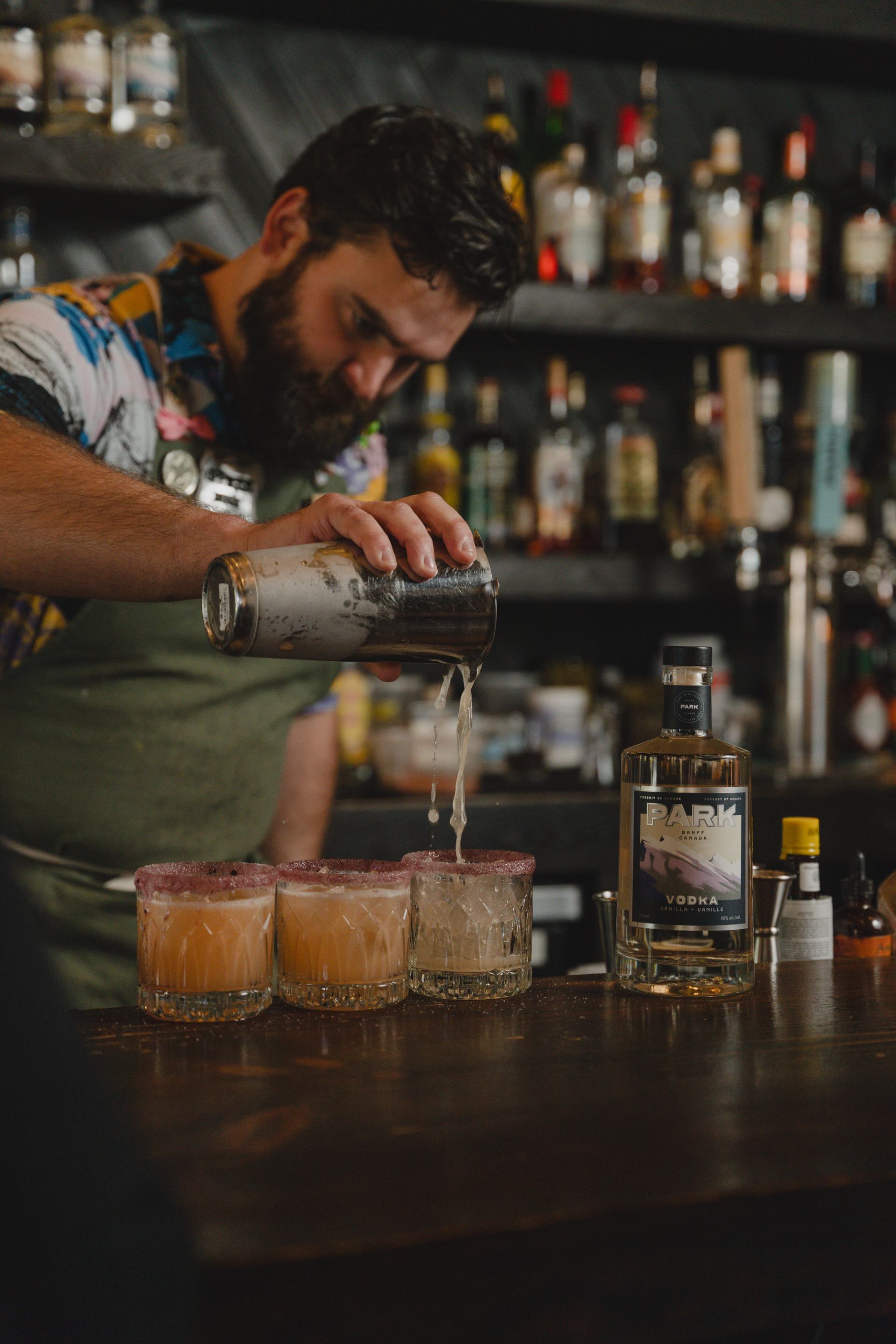 A bearded bartender pours a cocktail from a shaker into three rimmed glasses on a wooden bar.