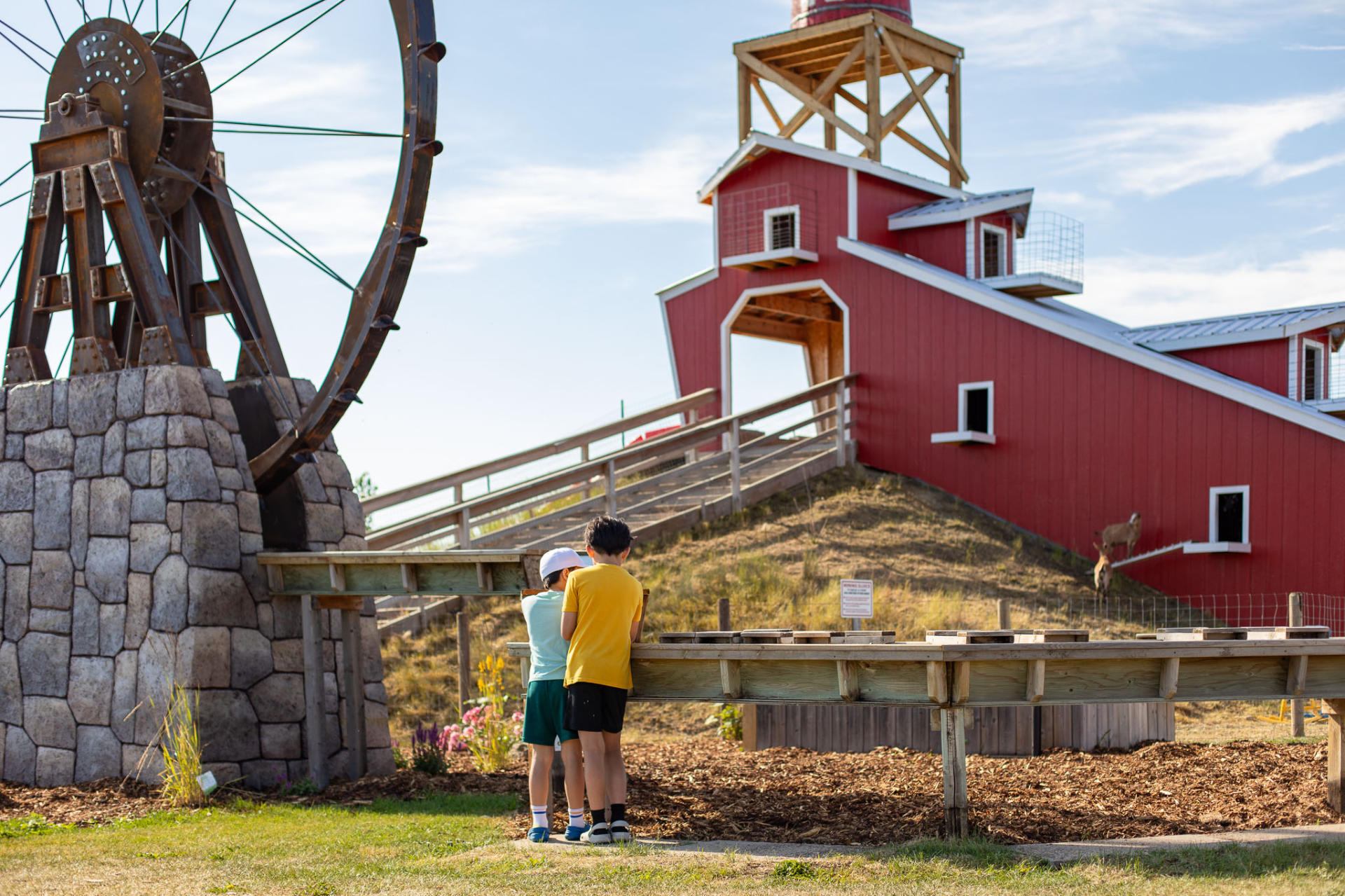 Children stand by wooden water feature near large wheel and red barn structure.