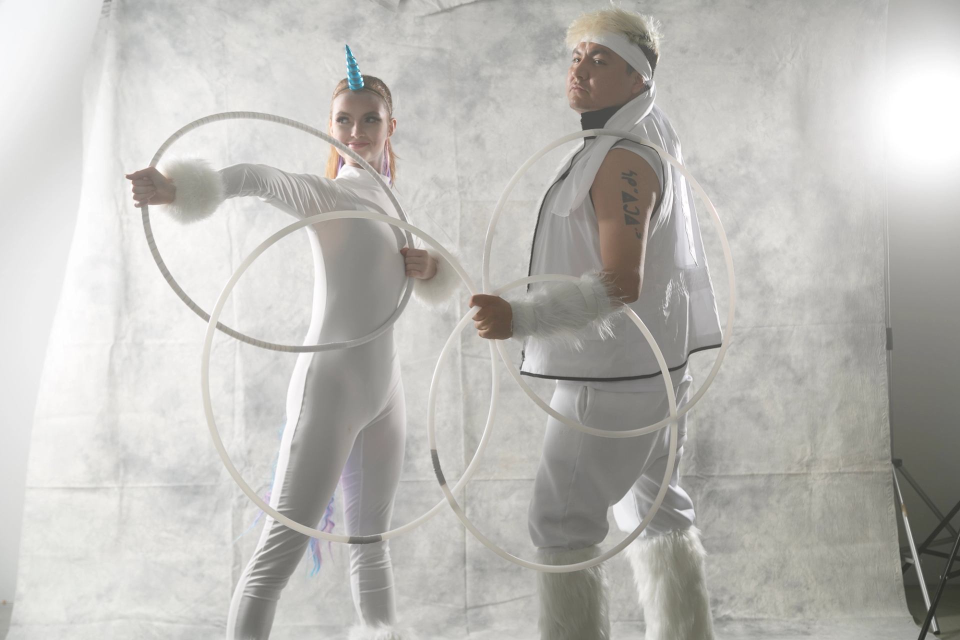 Two performers in white fantasy costumes pose with large hoops in studio lighting.