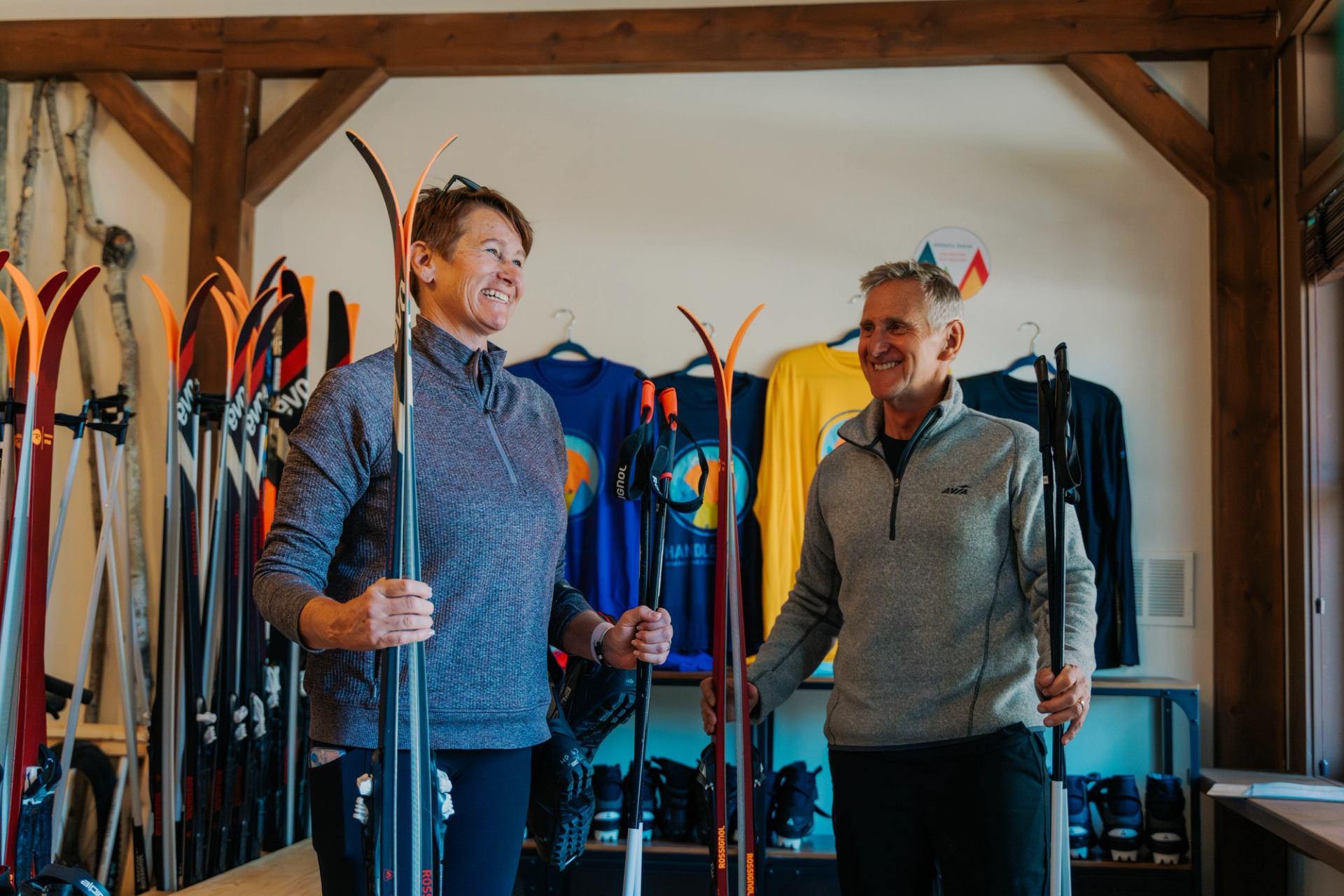 Two people holding ski poles inside a ski shop, with skis and jackets on racks.