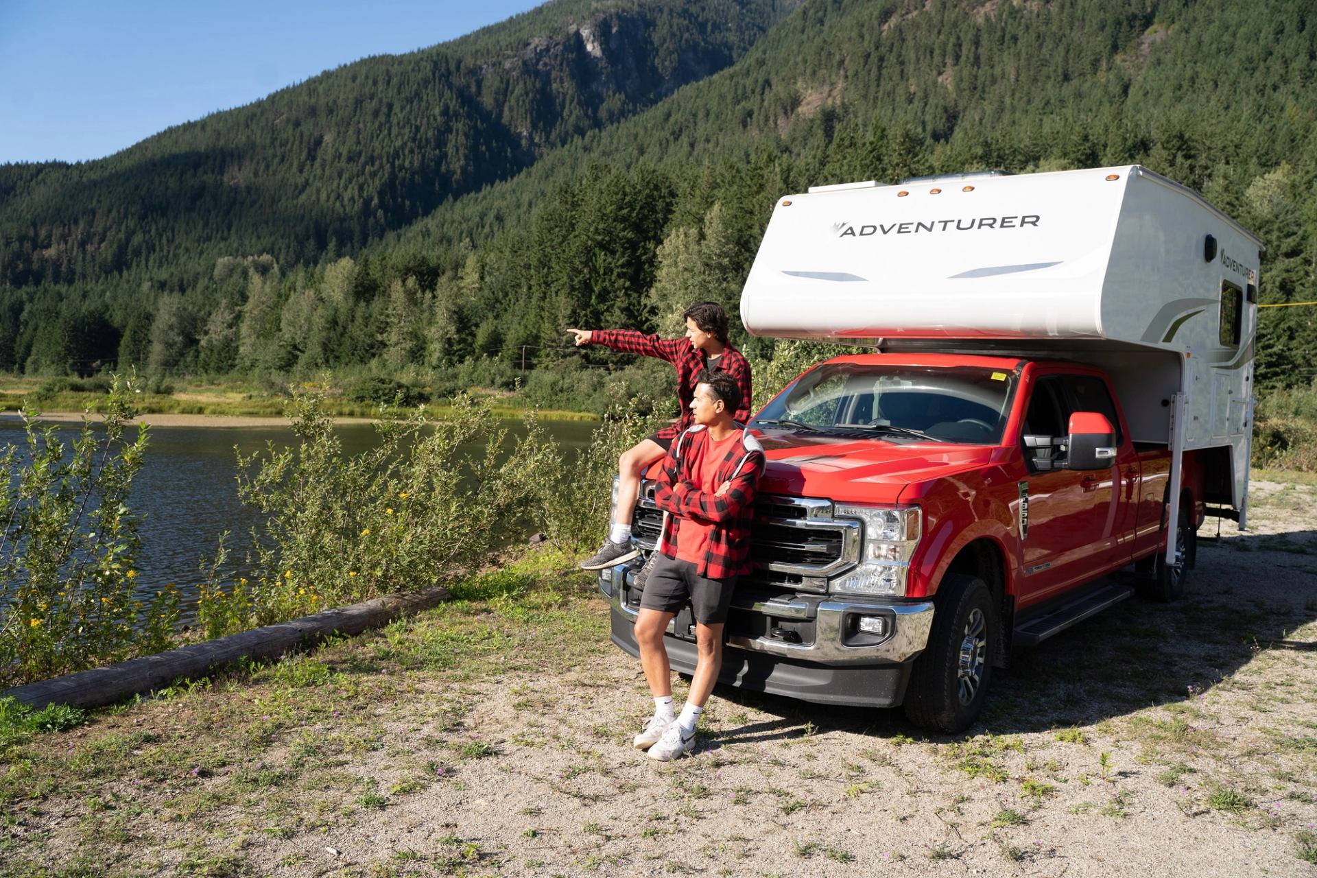 Red truck with camper parked by lake; two people enjoy mountain views.