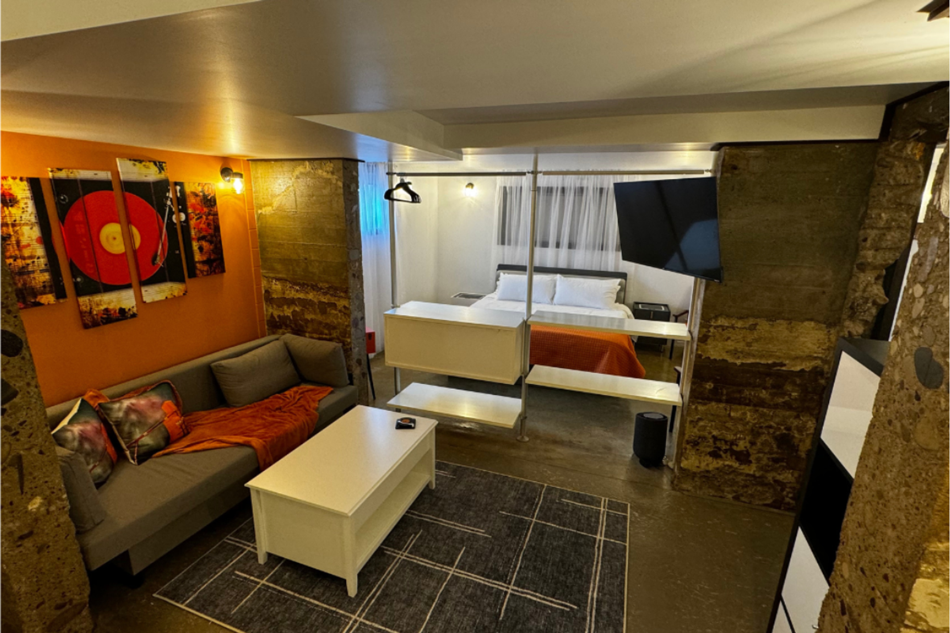 Open-concept room with grey sofa, white coffee table, bed and orange accents.