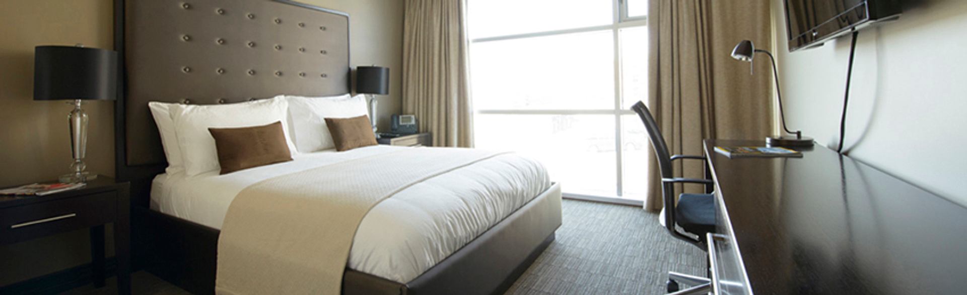 A modern guest room features a king bed, workspace, television, and large sunlit window with curtains.