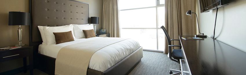 A modern guest room features a king bed, workspace, television, and large sunlit window with curtains.