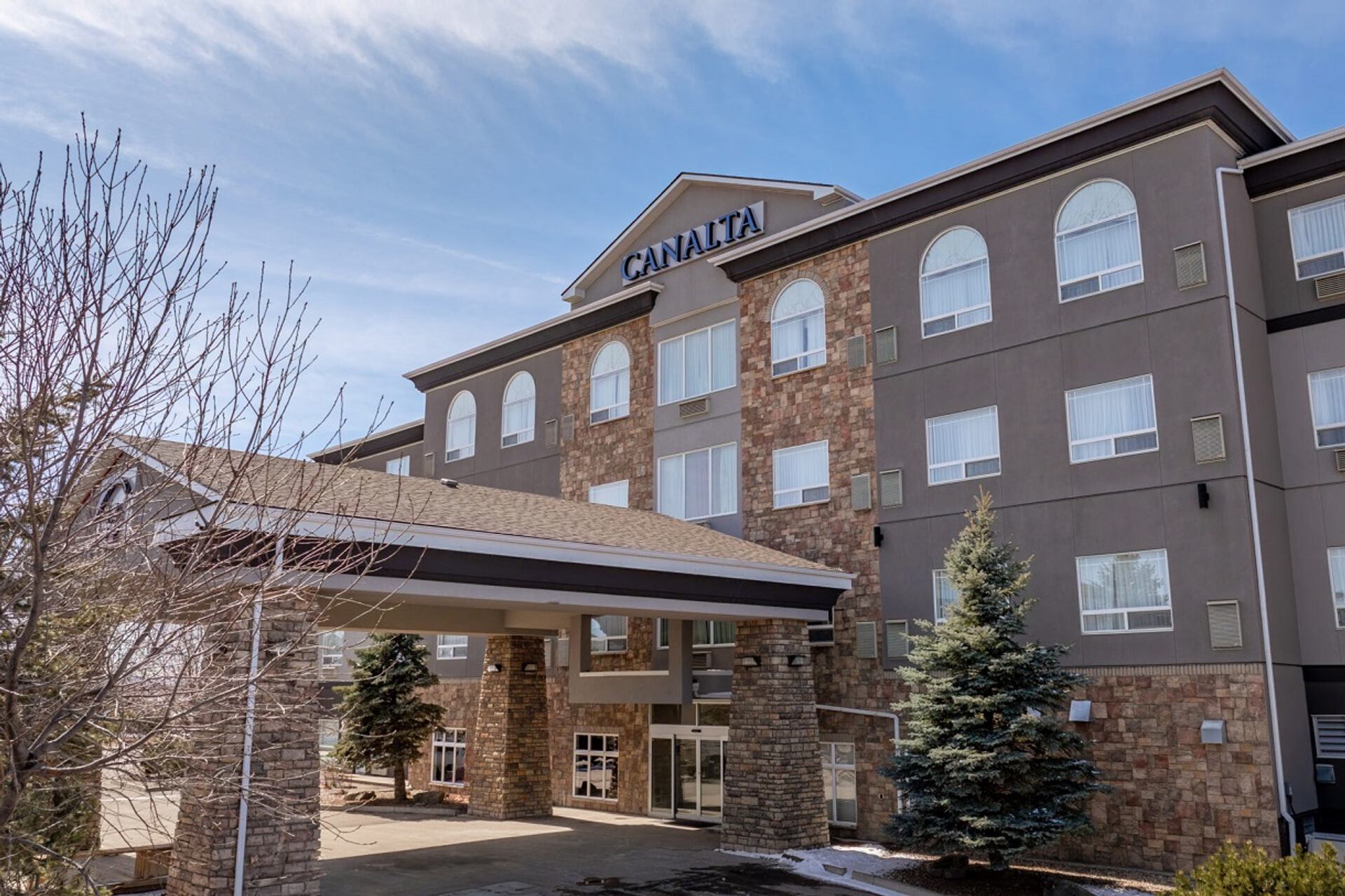 Canalta hotel exterior with stone facade, covered entrance and evergreen trees under blue sky