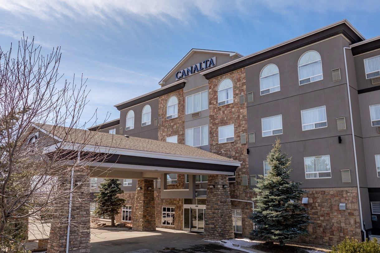 Canalta hotel exterior with stone facade, covered entrance and evergreen trees under blue sky