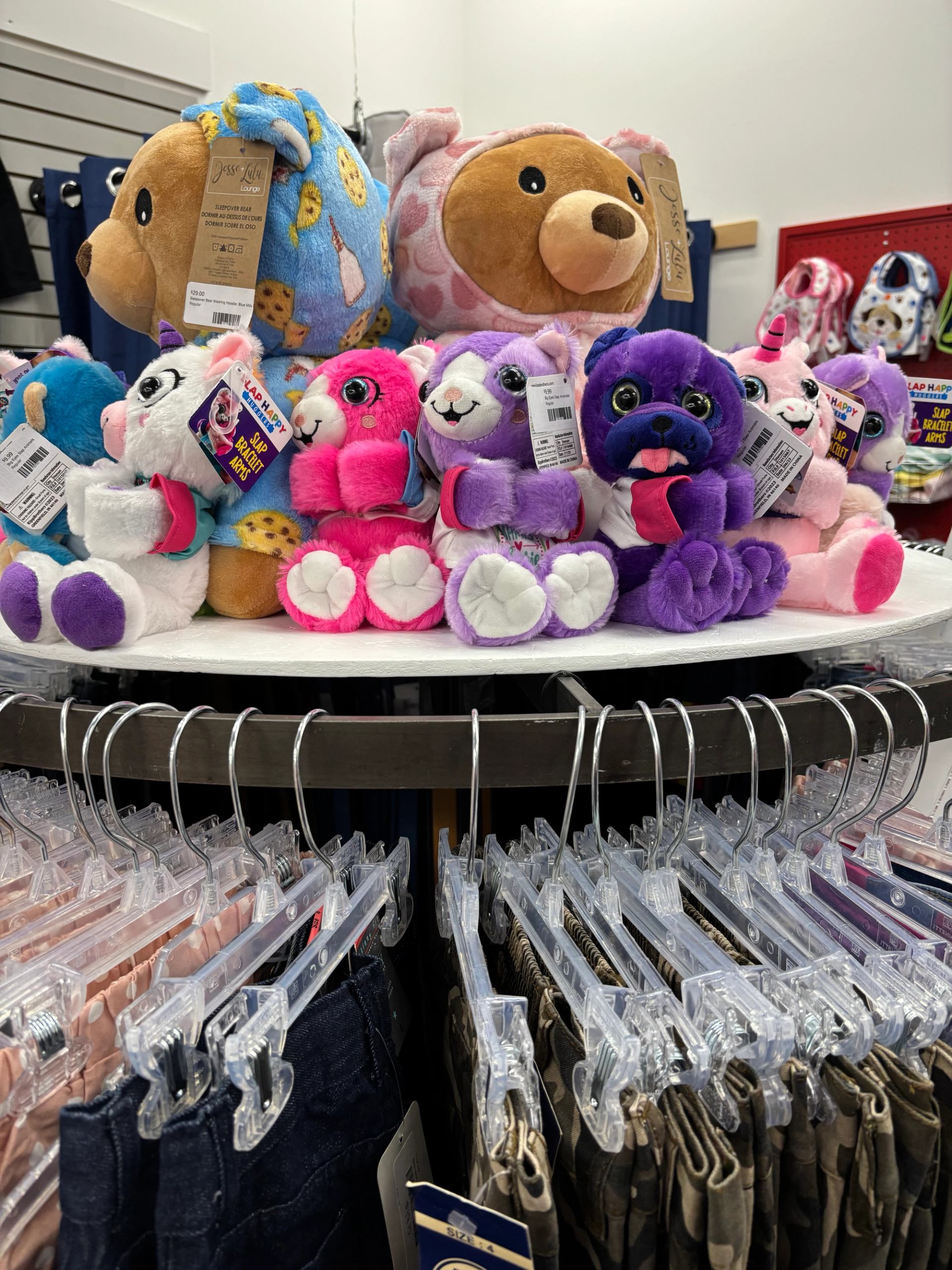 Display of colourful stuffed animals arranged on a circular rack above children’s clothing in a store.
