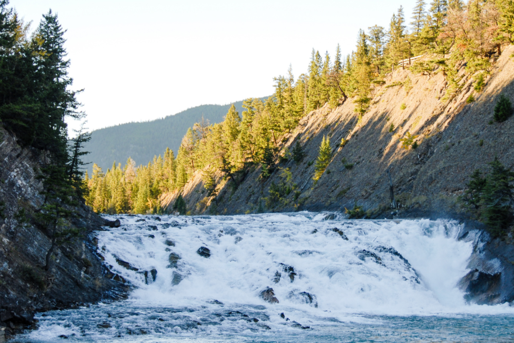 Wide waterfall flowing through a rocky canyon with forested hills in the background.