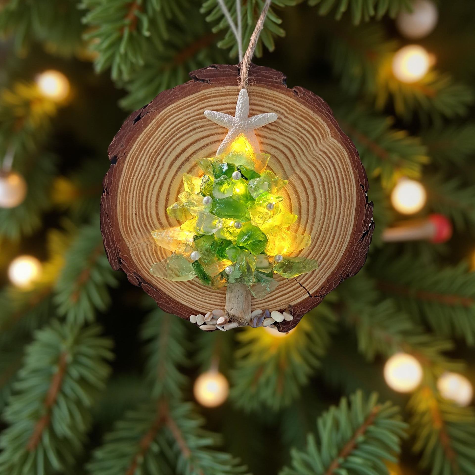 Wood slice ornament with glowing glass tree hangs on evergreen branches with lights.