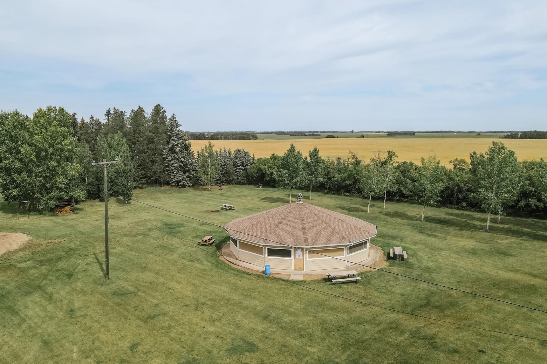 Octagonal pavilion on large grassy field with picnic tables and surrounding trees.