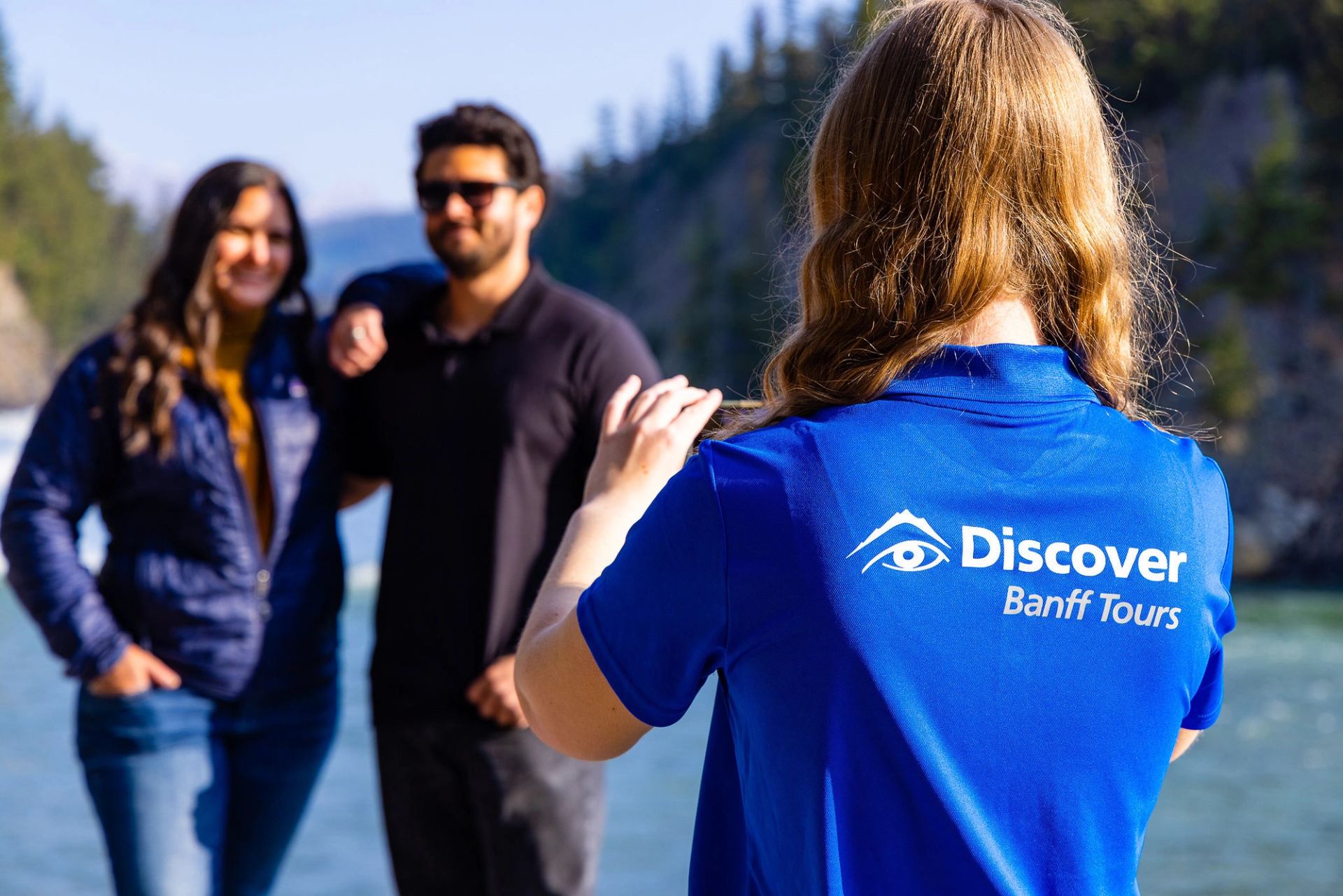 Tour guide in Discover Banff Tours shirt photographs two people by a river.