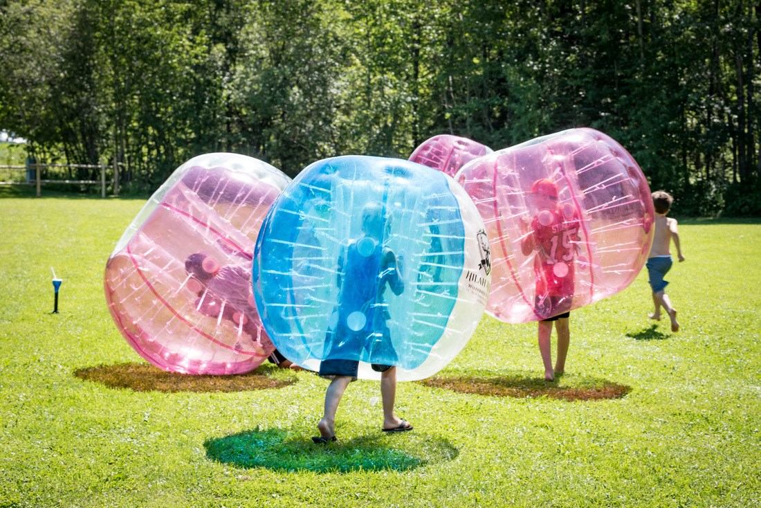 Children wearing large inflatable bubble balls play on a grassy field near trees.