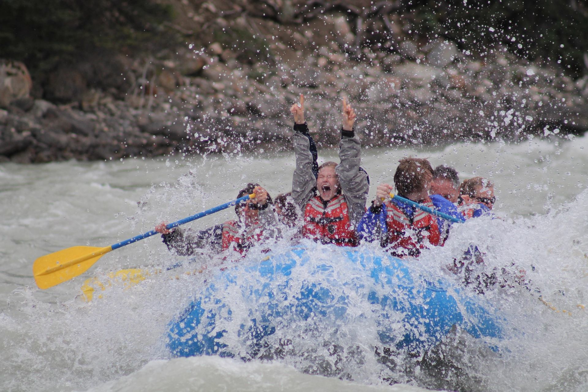 Rafters in life jackets celebrate with raised arms as waves crash over a blue raft in fast white water.