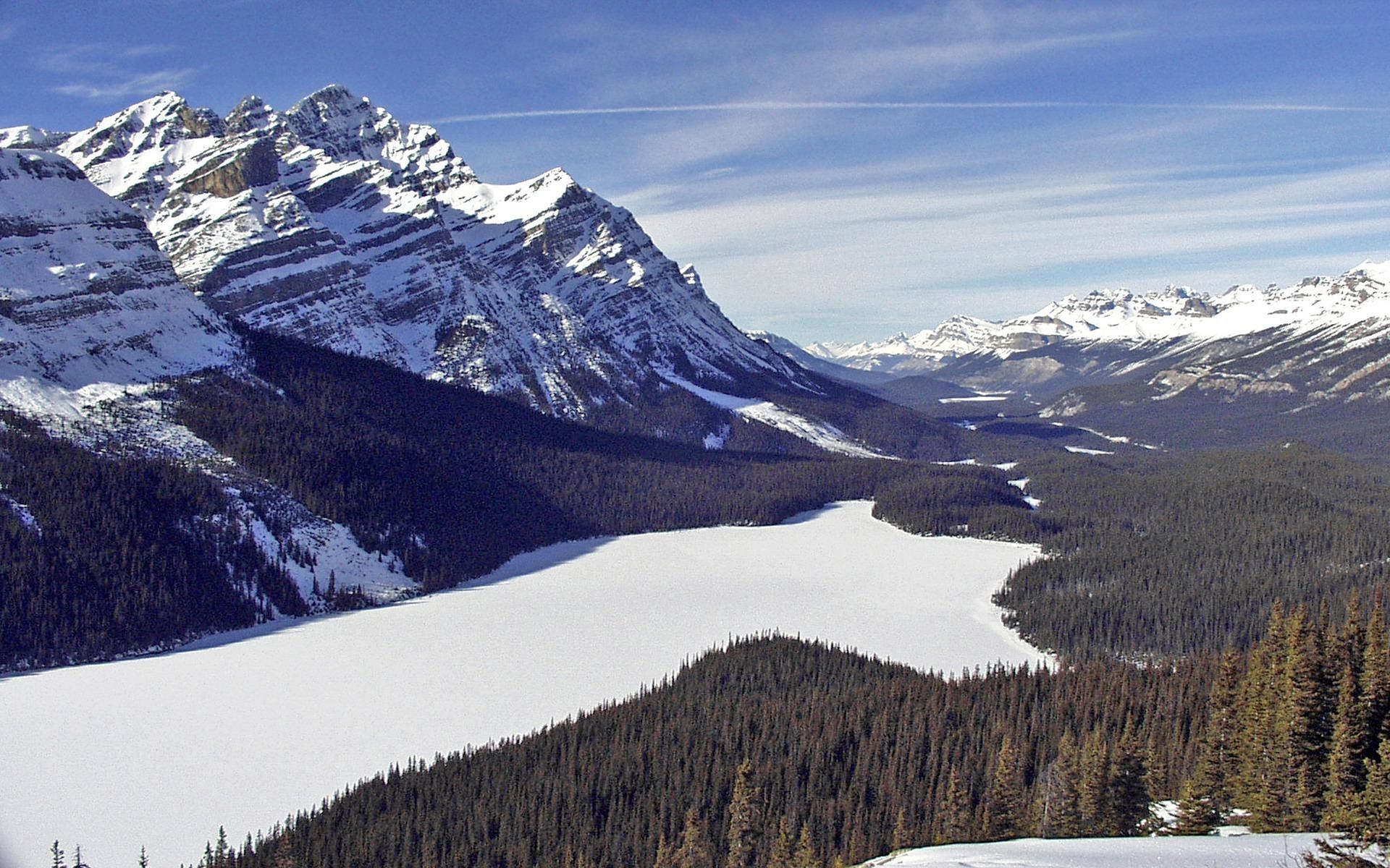 Snow-covered mountain peaks surround a frozen lake and dense evergreen forest.