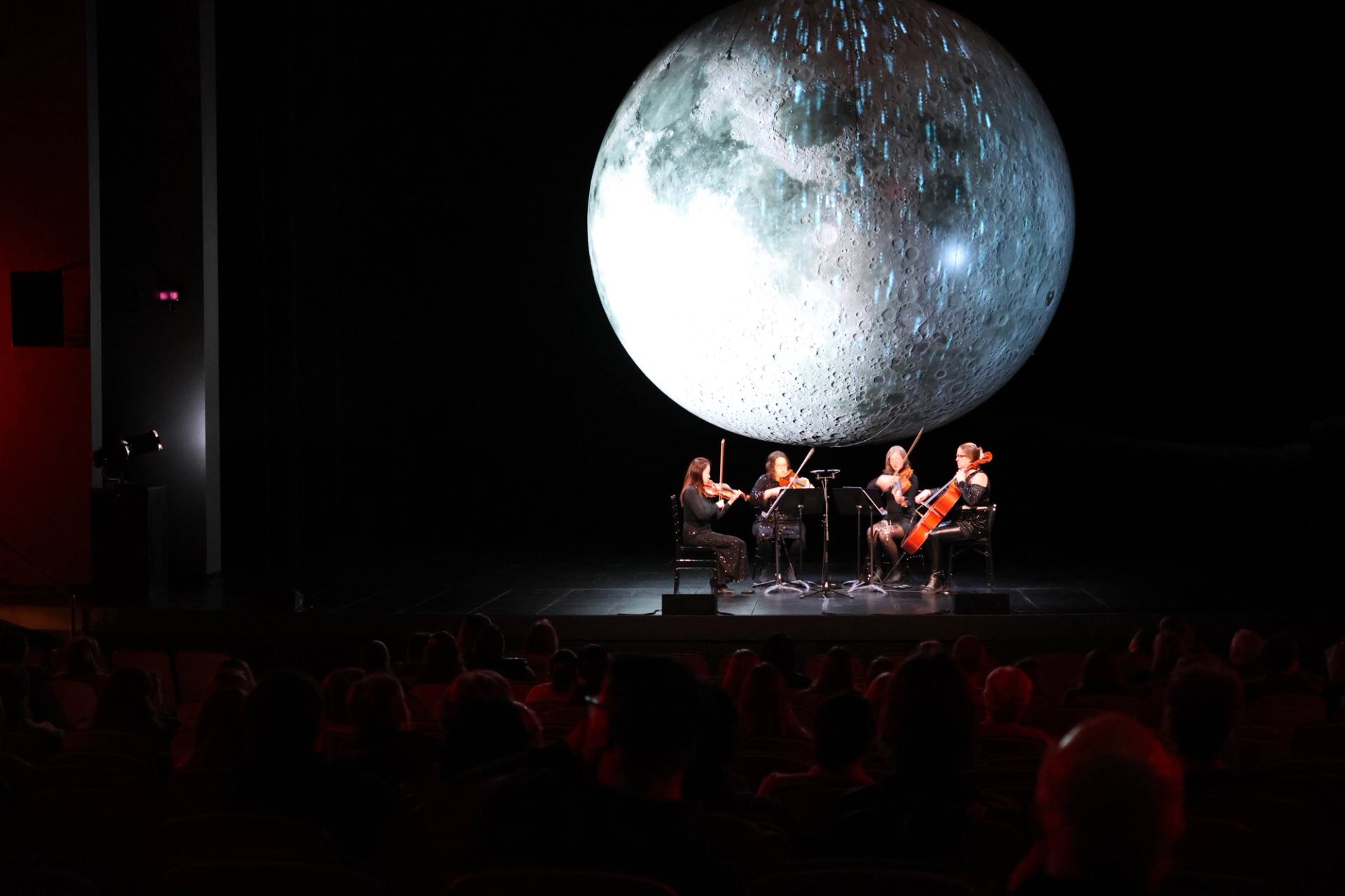 String quartet performs on dark stage before large illuminated moon backdrop, audience watching.